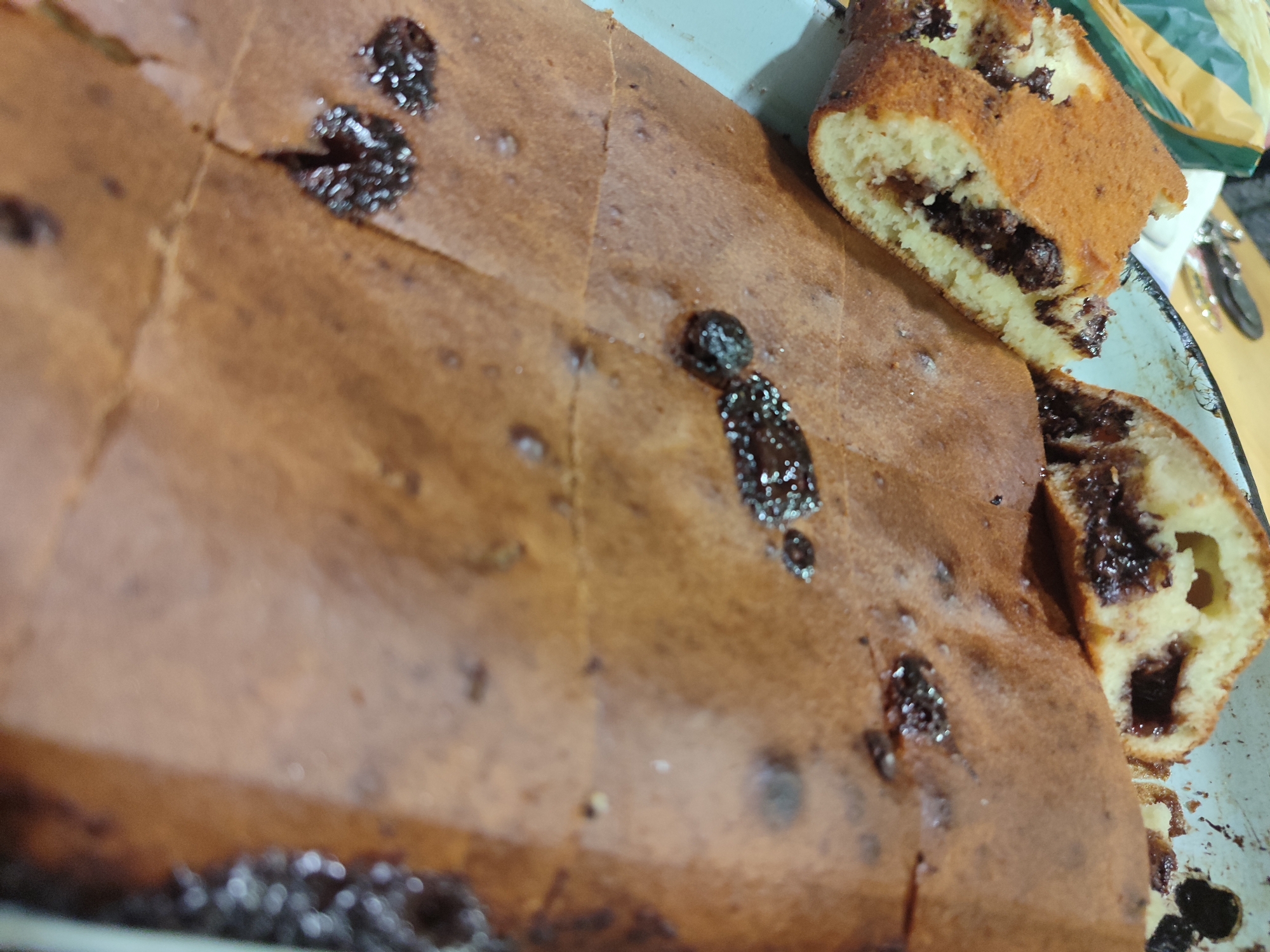 The image shows a freshly baked cake in a rectangular baking pan. The cake appears to be chocolate chip, with visible dark spots on the surface and a layer of chocolate filling visible in the cut slices. Several pieces have been cut from the cake, revealing the interior. The baking pan is light-colored. The cake and baking pan are positioned on a surface with other indistinct items nearby. The photo does not provide evidence to specify the location to be Varshilo, Bulgaria. No people are visible in the image. The environment includes a baking pan, a partially cut cake, and some objects out of focus in the background.