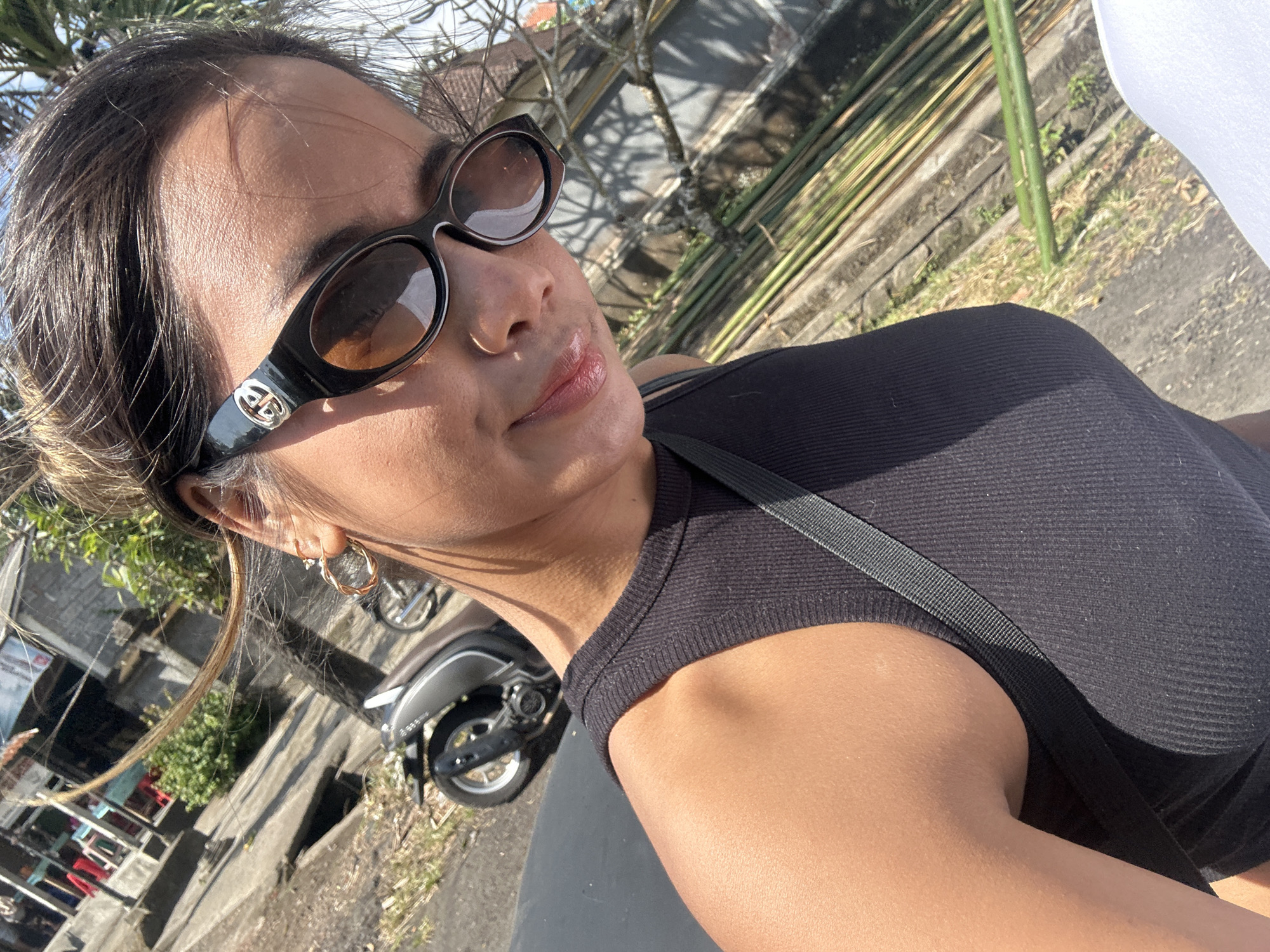 A woman wearing sunglasses and a black tank top takes a selfie in a street in Sukawati, Indonesia. She has a gold hoop earring in her left ear. Behind her, a motorcycle is parked near a building. The street is paved and there are plants and bamboo stalks in the background. The sun is shining.