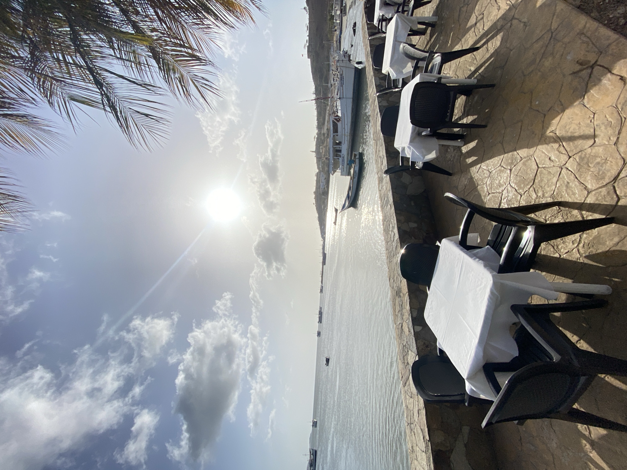The image depicts a restaurant patio with black tables and chairs, each table covered with a white tablecloth, situated along the waterfront in Juan Griego, Venezuela. The patio surface appears to be made of interlocking paving stones. Palm fronds are visible in the upper left corner. The ocean stretches into the distance, dotted with boats. The sun is high in the sky, partially obscured by clouds, casting a bright glare on the water. Across the bay, a hillside or townscape is visible.