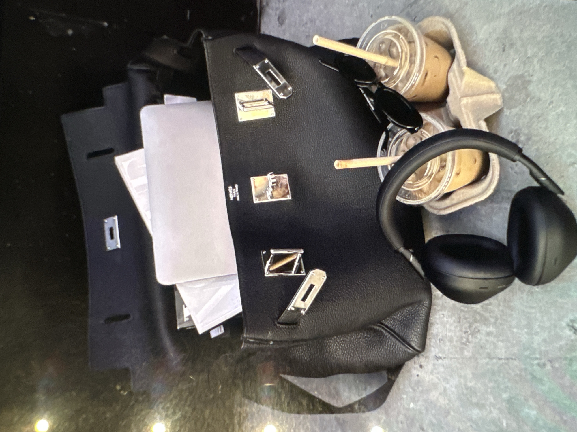 The image depicts a black leather handbag with silver hardware open, revealing a laptop and papers inside. Next to the bag are two iced coffee drinks in a cardboard carrier, a pair of black sunglasses, and black over-ear headphones. The scene is likely outdoors, with a slightly textured gray surface beneath the objects. The city and country are not discernible from the image.