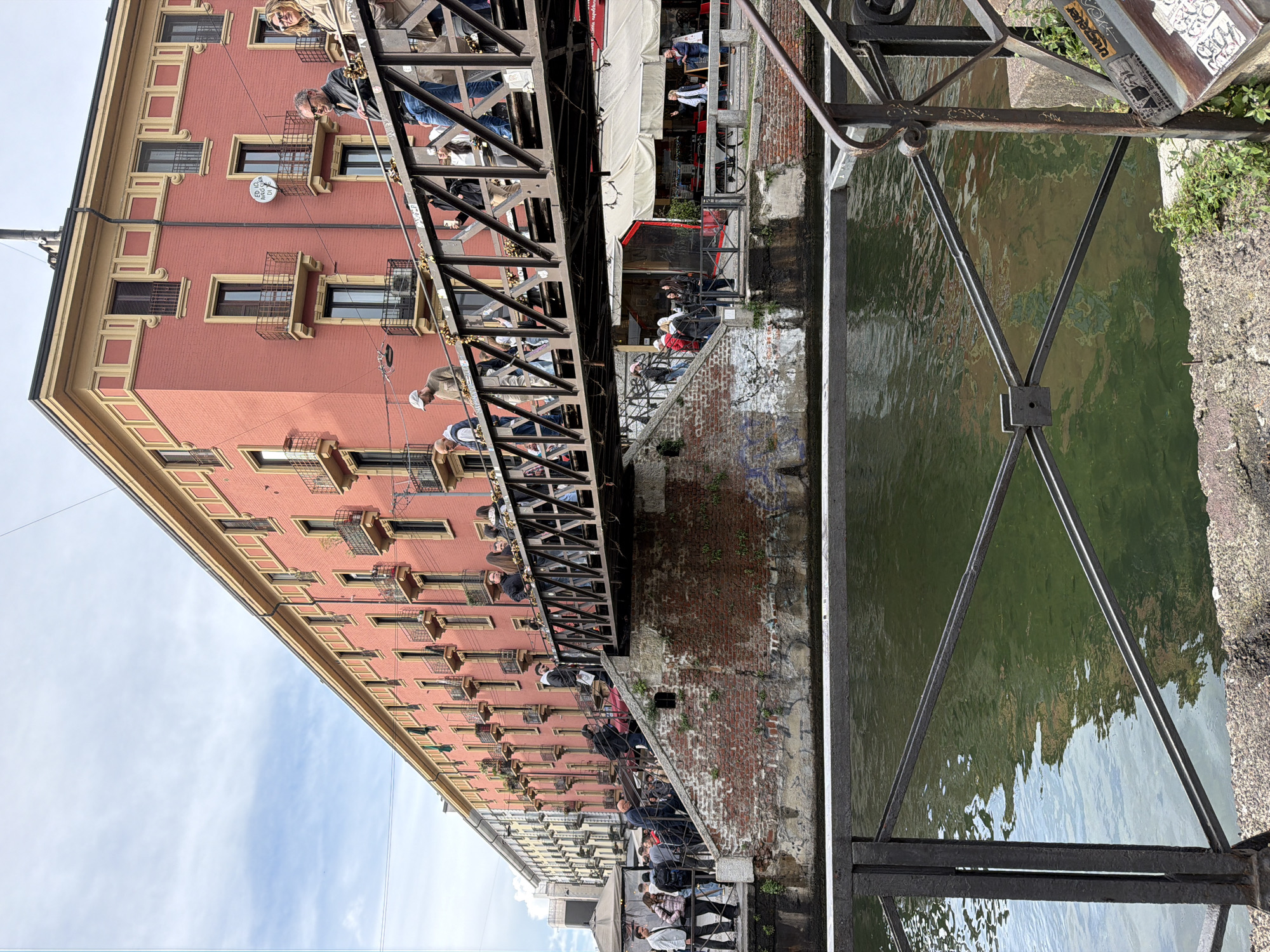 The image depicts a canal scene in Milan, Italy. The canal water is green and reflects the sky and surrounding buildings. A metal pedestrian bridge crosses the canal. People are standing on the bridge, looking at the canal and surrounding area. A red brick building with yellow trim lines the canal. The building has many windows with small iron balconies. Below the bridge, people are walking along the canal bank. A white canopy covers a section of the canal bank. The sky is blue with some clouds. A metal bridge support structure is visible on the right side of the image. The perspective is unusual, making it appear as if the image is rotated counterclockwise.