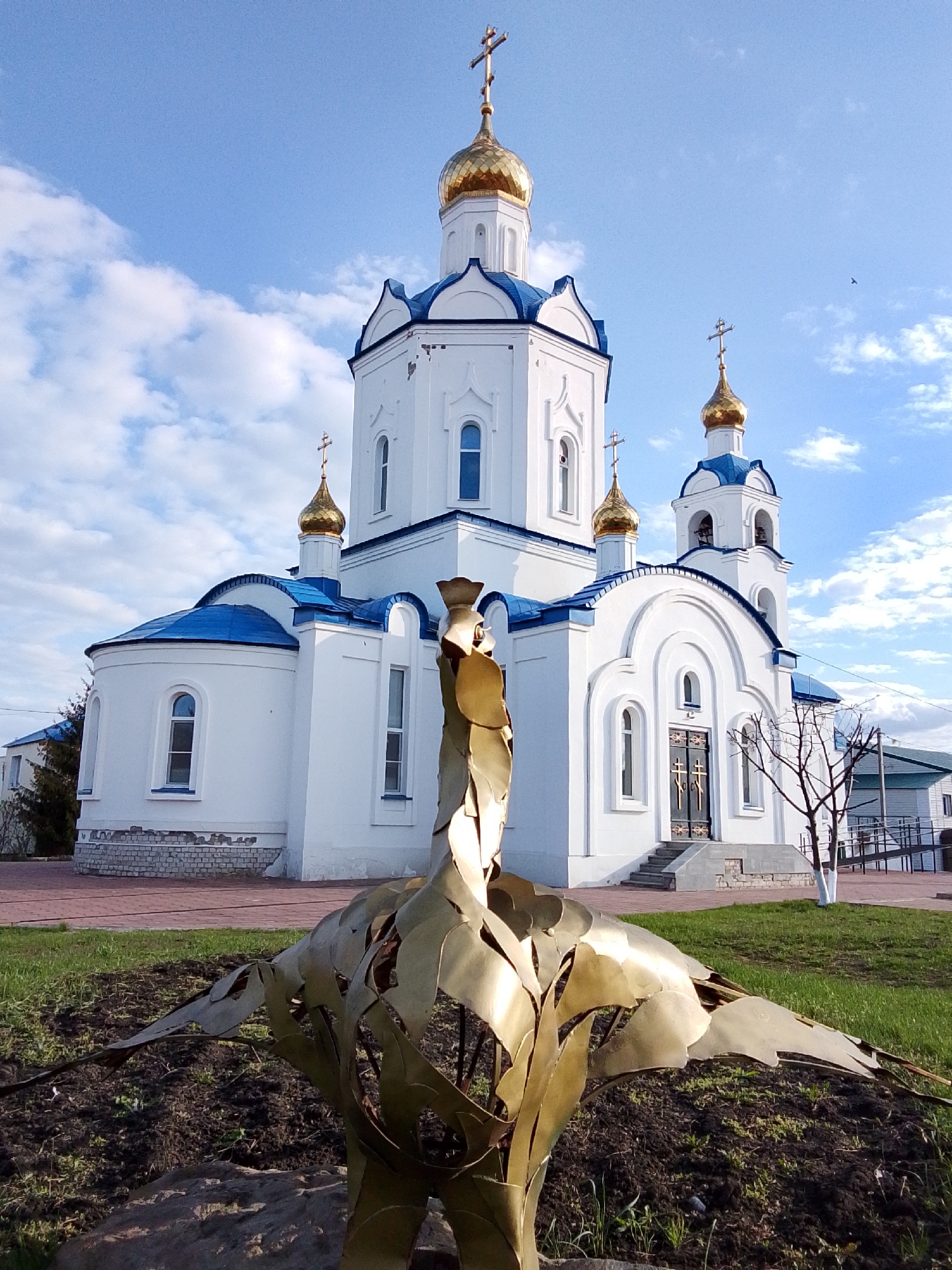 The image displays a prominent white Orthodox church with multiple golden onion domes and distinct blue roofs, situated under a bright blue sky dotted with scattered white clouds. The church features arched windows and doorways, and several golden crosses crown its domes.
In the immediate foreground, a large, golden, abstract sculpture resembling a stylized bird with broad, textured wings is planted in a patch of dark soil. The metallic surface of the sculpture gleams, catching the sunlight.
The church is surrounded by a lawn of green grass and some bare soil. To the right of the church's main entrance, a small, leafless tree stands. Paved areas are visible around the church, and other buildings, including one with a ramp, are partially visible in the background. The scene depicts a serene outdoor environment, likely a churchyard or public square in Khlevnoye, Russia. The time of day appears to be sunny daytime, possibly late afternoon or early morning, given the light quality. No people or specific events are captured in the photograph, and there is no visible text. A small bird is visible flying in the distant sky.