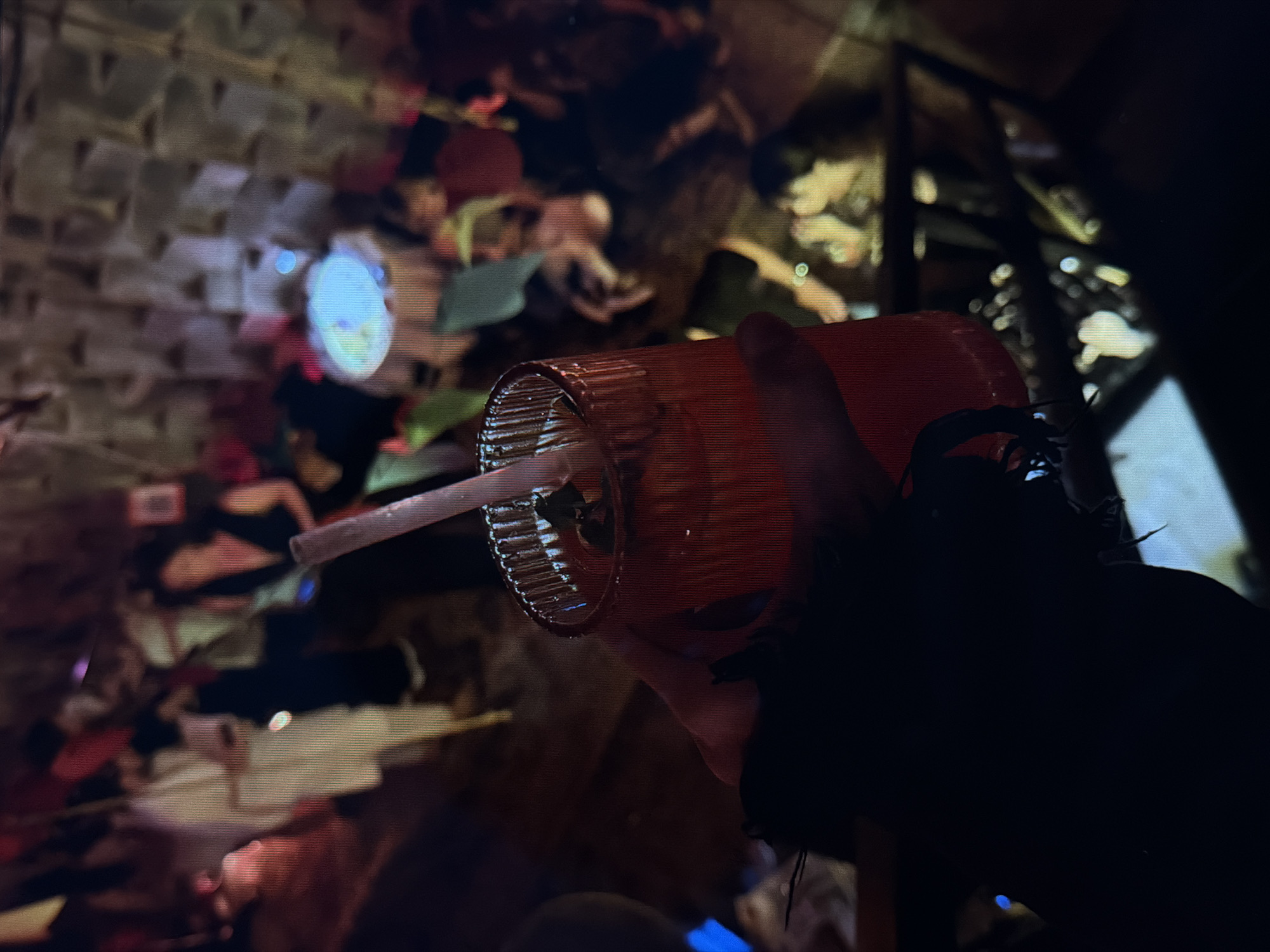 The image depicts an illuminated red-orange beverage glass, featuring vertical ribbing, containing a straw. The glass is held by a partially visible hand. A dark, unidentifiable object is positioned to the right of the glass. The background is blurred, but suggests a busy urban environment, possibly a night market or street food area in Đà Nẵng, Vietnam. This assessment is based on the presence of indistinct figures, indistinct structures and lighting consistent with such a scene.