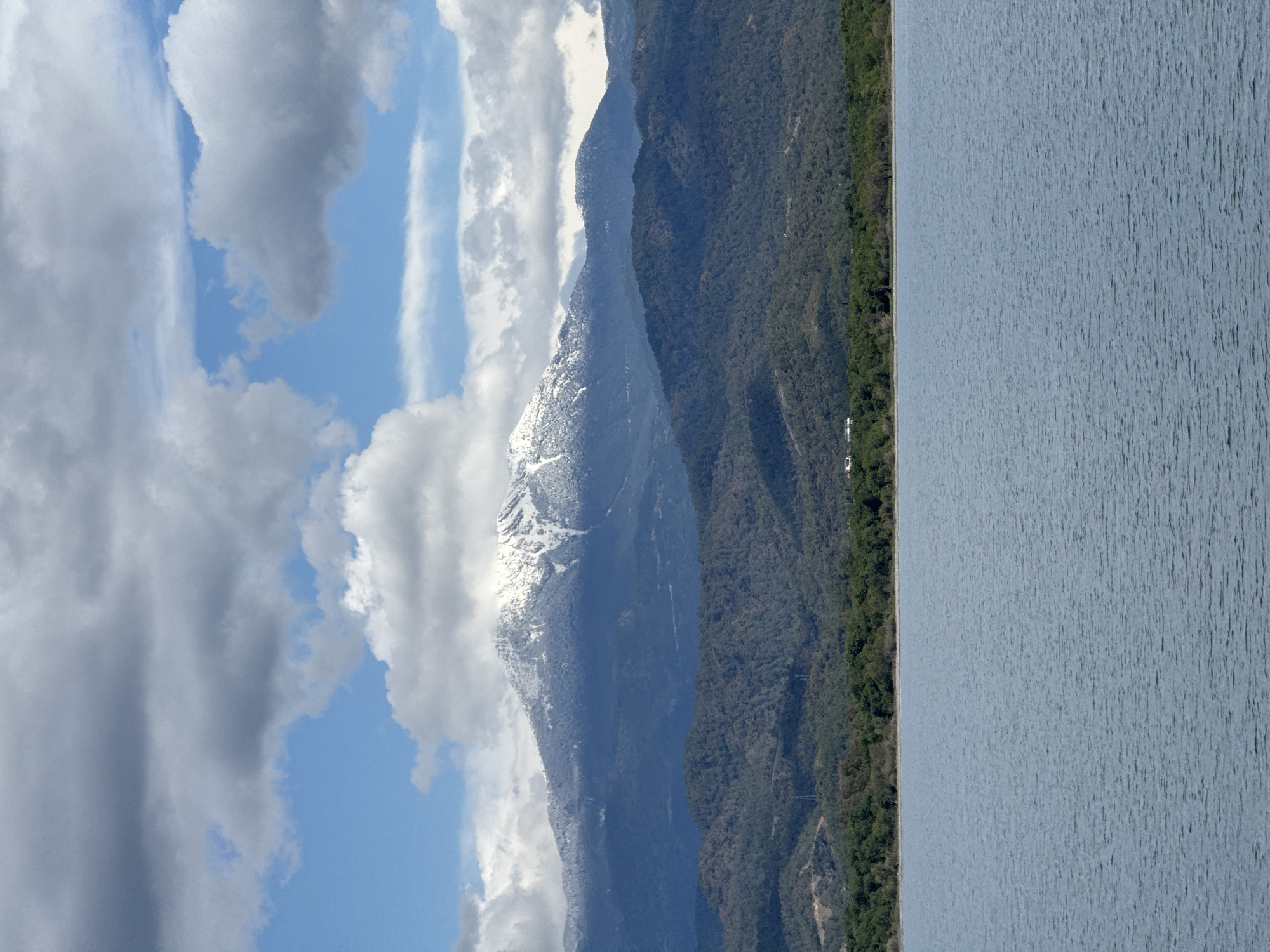 The image depicts a landscape in an unknown location in Türkiye, featuring a body of water on the right side, and mountain ranges on the left. The mountain closest to the viewer is covered in snow towards its peak. Forests cover the lower slopes of the mountains. The sky is visible in the upper-left, featuring blue patches and varied cloud formations. The image is rotated 90 degrees counterclockwise.