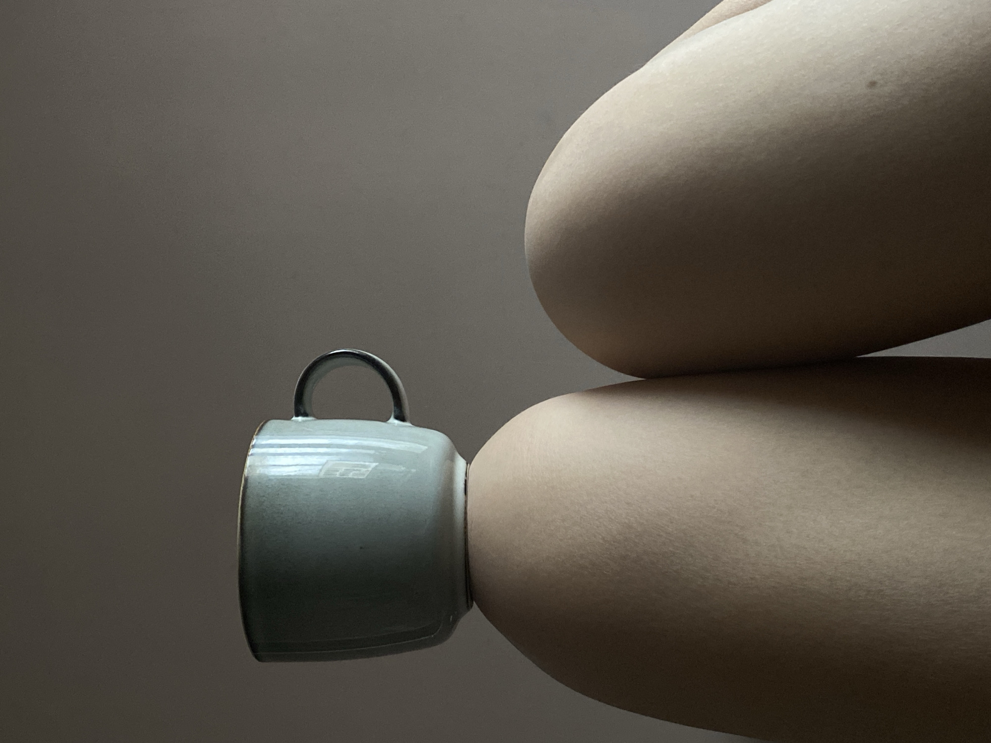 The image presents a close-up, abstract composition featuring a light grey ceramic mug nestled between two smooth, curved sections of human skin, likely an arm or leg. The mug, positioned horizontally, appears to be gently squeezed or held in place by the body parts, which form soft, rounded shapes above and below it. The upper body part casts a subtle shadow on the lower one, and a small, dark mole is visible on the surface of one of the skin sections, particularly evident in one of the cropped hints.
The background is a plain, uniform dark grey wall, providing a stark contrast to the subjects and emphasizing their textures and forms. Soft, diffused lighting illuminates the scene, highlighting the subtle contours and smooth surface of the skin and the slightly reflective glaze of the mug. No specific time of day or outdoor weather conditions can be discerned. The overall color palette is muted, consisting of warm beige skin tones, cool grey of the mug, and the neutral dark grey of the background. There is no visible text in the image.