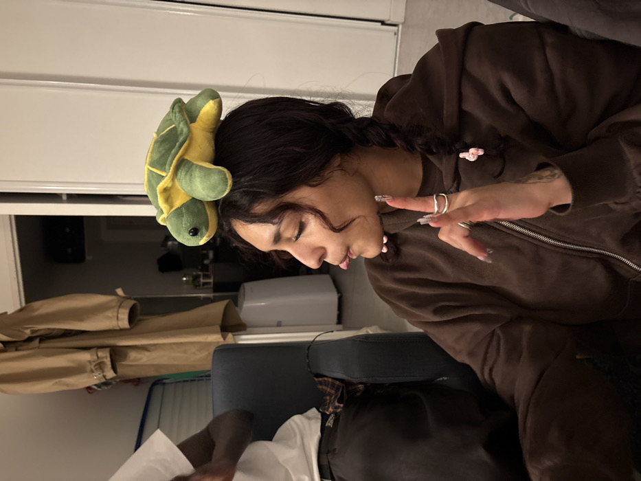 The image depicts an indoor scene with two individuals. In the foreground, a female with dark, curly hair in braids is seen in profile, facing left. A plush green and yellow turtle toy is balanced on the crown of her head. She wears a brown zippered hooded jacket, has multiple rings on her left hand, and her fingernails are painted. Her left hand is raised, with the index finger extended upwards. Her tongue is slightly extended from her mouth. A tattoo is visible on her left forearm. To her left, partially visible, is another individual with a darker skin tone wearing a white t-shirt and seated on a dark blue armchair. A plaid garment is discernible near this individual's lap. The background shows a neutral-toned wall or door frame. A beige trench coat hangs on a hook on the left side of the background. A white, tall, cylindrical appliance, possibly an air purifier or humidifier, stands in the mid-background. The specific location within Paris, France, cannot be determined from the visual information provided.