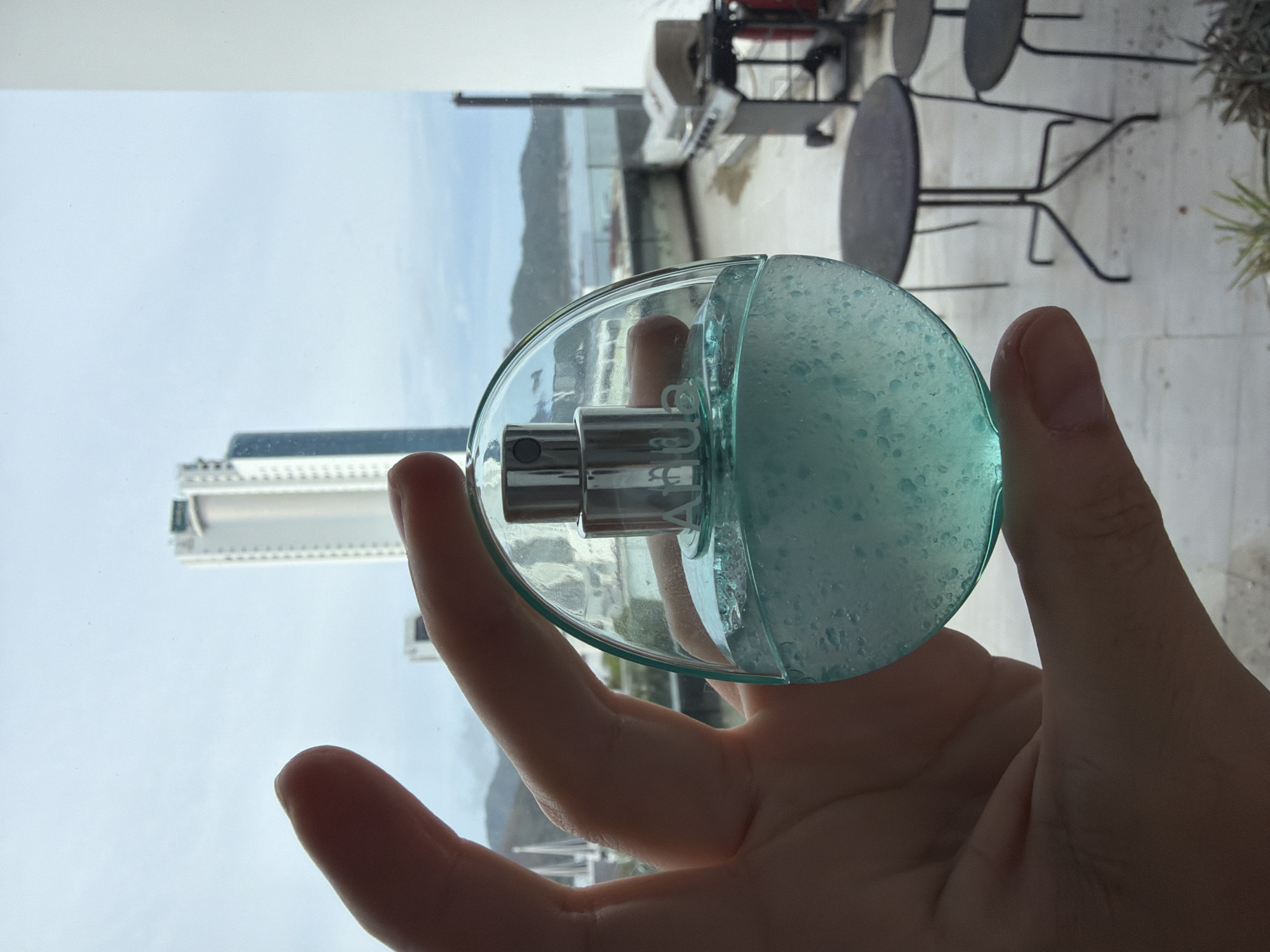 A hand, predominantly in silhouette, holds a transparent, circular glass bottle against a bright, distant city and coastal backdrop. The bottle is partly filled with a pale blue, bubbly liquid and features a metallic spray mechanism at its top.
The setting appears to be a high-rise balcony or rooftop overlooking a coastal city, likely Bac Nha Trang Ward, Vietnam. In the background, a wide expanse of a light blue sky dominates the upper portion. Below the sky, the landscape features distant mountains or hills meeting a body of water, indicative of a sea or ocean. Several modern, tall buildings are visible in the cityscape, with one distinct white high-rise prominent on the left.
To the right of the frame, parts of an outdoor seating area are visible, including two dark, round tables and chairs, suggesting a patio or restaurant area. The time of day is daytime, with a clear or lightly overcast sky, creating a slight silhouette effect on the foreground elements.
On the metallic part of the bottle's dispenser, mirrored text is faintly visible, which appears to spell "HUG". The hand is holding the bottle by its base, with fingers curled around its circular shape.
