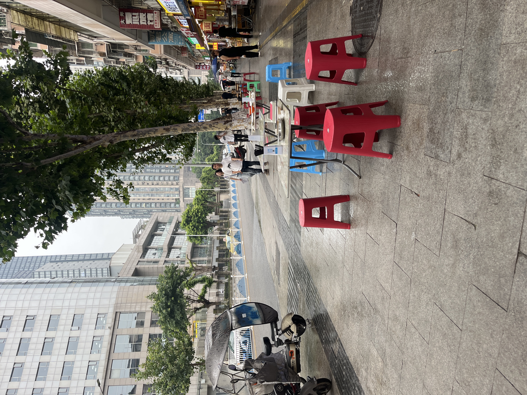 This is a street scene in Yuzhong District, China, likely in the morning or late afternoon given the overcast sky and street lights. The foreground is dominated by a wet, gray tiled sidewalk, indicating recent rain. Several red plastic stools are scattered on the sidewalk, some positioned around small tables set up for dining. A motorized three-wheeled vehicle with a protective canopy is parked on the left, also appearing wet. Beyond the tables, a street runs parallel to the sidewalk, with a white and blue barrier separating the pedestrian area from the road. Cars are visible on the road.
On the right side of the street, a row of buildings with shops at ground level is visible. These buildings have multiple floors and many windows. Some of the shops have awnings and display signs with Chinese characters. People are walking along the sidewalk and in front of the shops. A few individuals are interacting at the tables, suggesting an outdoor dining area for one of the establishments. The overall atmosphere is that of a typical urban street in China, with signs of commercial activity and pedestrian life, potentially a food street or market area. The tall modern buildings in the background contrast with the older, more densely packed buildings on the street level.