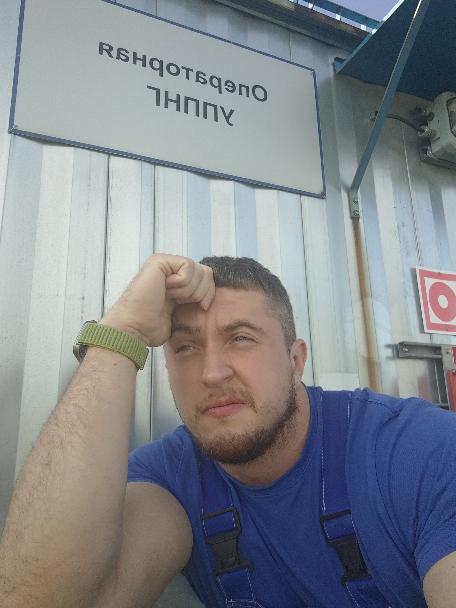 A man in a blue shirt and overalls with a green watch on his left wrist rests his forehead on his left hand, looking to his left. He has a beard and short, dark hair. He is in front of a corrugated metal wall. Above him, a white sign with dark text in a foreign language is mounted on a blue frame. The sign's text is illegible in the original image. To the right of the man, a red and white warning sign with a circular red symbol is visible. The overall environment suggests an industrial or construction setting.