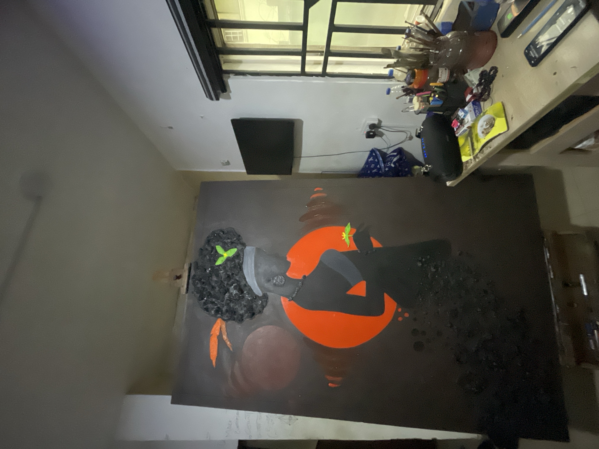 The image shows a painting leaning against a wall in an indoor setting. The painting depicts a profile of a person with afro hair, adorned with two leaves, set against an orange circular shape. The background of the painting is black, with some areas showing textured patterns and a brown circular shape.
Near the painting, there is a window and a table with various objects, including what appears to be art supplies such as brushes and paint. A black rectangular object is mounted on the wall next to the window.
Without any additional context or identifying features, it is not possible to confirm the location as Ado, Nigeria.