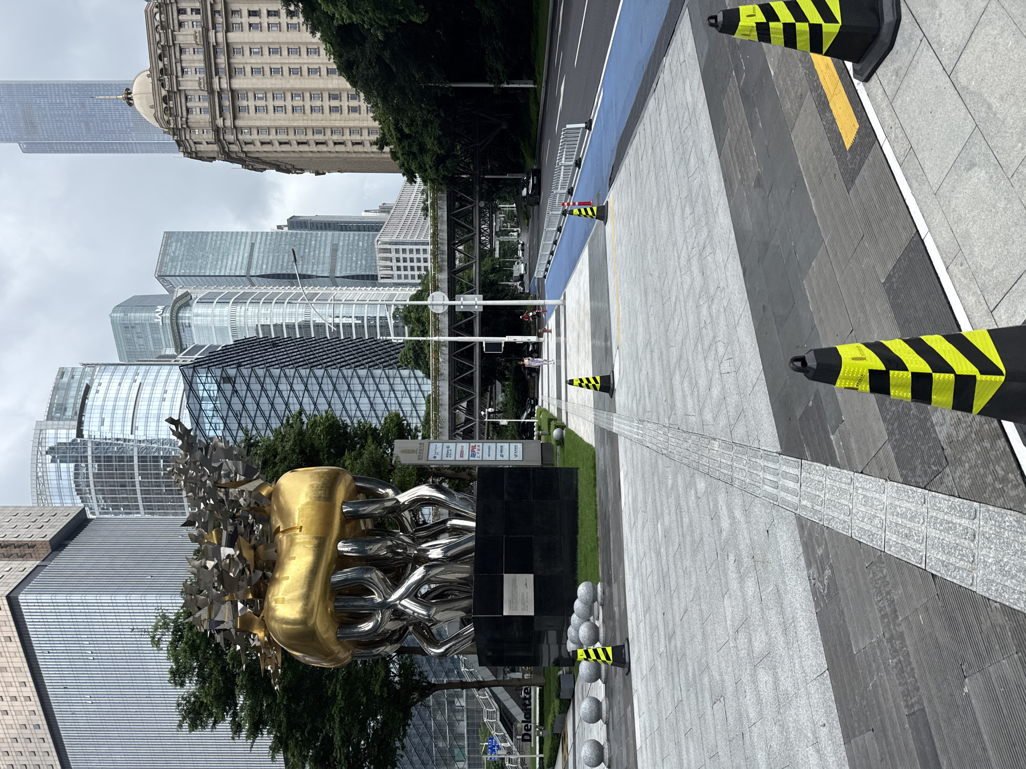 This is a street-level, medium-shot photograph taken in Tianhe District, China. The scene captures a modern urban environment with a focus on a large, abstract sculpture in the foreground. The sculpture is predominantly gold and metallic, with a bulbous top and intricate metallic supports. It is situated on a dark, rectangular base.
To the left of the sculpture, several round, granite spheres are arranged in a line. The ground is paved with large, gray tiles, and a tactile paving strip runs along the path. Black and yellow traffic cones are placed strategically on the pavement, indicating a construction or safety zone.
In the background, a series of modern skyscrapers with glass facades dominate the skyline. A pedestrian overpass crosses the street in the mid-ground. The street itself features a blue bike lane, separated from the main road by a white metal barrier. Several cars are visible on the road.
To the right, a large, older building with a domed roof and classical architectural details stands out against the modern skyscrapers. Lush green trees line the street and parts of the background, providing a contrast to the concrete and glass structures. The sky is overcast, suggesting a cloudy day.
There are a few individuals visible: one person walking on the left sidewalk and another person in red further back on the left side. Two people are visible in the mid-ground, walking on the overpass. The overall impression is one of a busy, contemporary city with a mix of old and new architecture, and public art.
