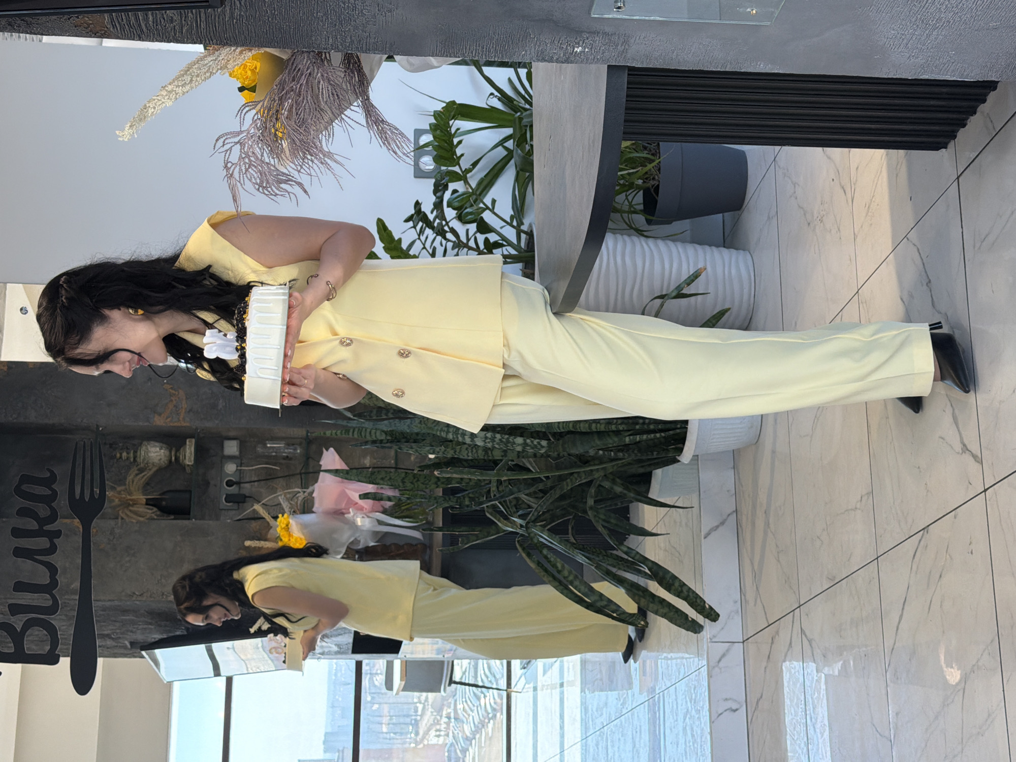 A female subject is depicted standing indoors on a white marble-patterned tiled floor in what is identified as "Ресторан Вилка" (Restaurant Fork) at Borodina St., 25A, Izhevsk, Russia. The subject has long, dark, wavy hair and wears a pale yellow sleeveless blazer-style top with gold buttons and matching wide-leg trousers, paired with black pointed-toe flat shoes. She is smiling faintly and looking towards her left. Both hands support a small, round white cake, decorated with dark toppings, gold sprinkles, and white candles forming the number "28".
The immediate background on the left features large windows providing a panoramic view of Izhevsk, revealing a cityscape and a parking area below, indicating an elevated floor. A large mirror reflects the subject and the exterior view. To the right of the mirror, a dark textured wall displays a prominent black silhouette of a fork and the Cyrillic word "Вилка". Below this sign, decorative items and potted plants are visible. Further to the right, a dark circular-topped table is partially visible, along with a bouquet containing yellow flowers and dried grasses, and several potted plants, including large snake plants in white and gray planters. The environment is well-lit, suggesting natural daylight.