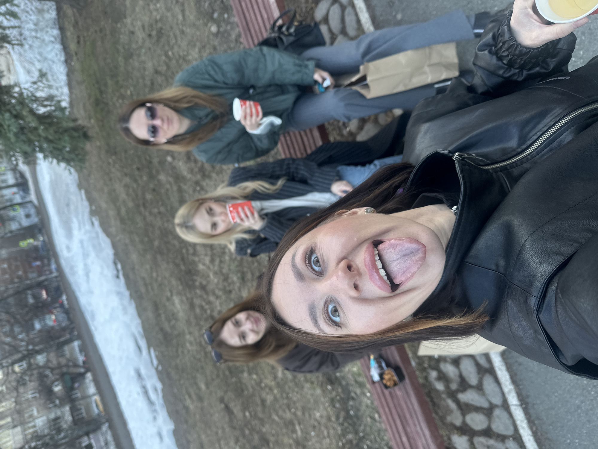 The image is a selfie of four young women sitting on a park bench in Izhevsk, Russia. The woman in the foreground is sticking her tongue out. The women in the middle each hold a red and white cup. The woman at the back wears sunglasses. The bench has a slatted seat and is situated on a patch of dry grass. There is snow on the ground in the background and a row of buildings is visible in the distance.