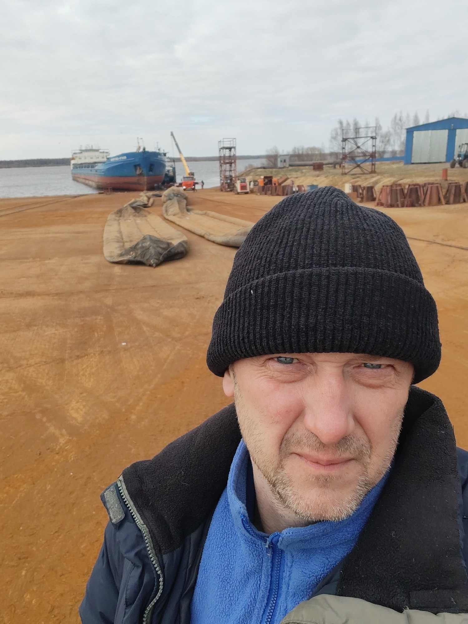 A man in a black knit cap and a blue fleece jacket is taking a selfie. He has fair skin, blue eyes, and short grey stubble on his face. The background shows an industrial riverside scene, likely a shipyard. A blue and red cargo ship is docked at the shore, with a crane nearby. On the land, there are large, heavy-duty fabric sleeves, possibly for lifting or moving heavy objects. Scattered around are metal structures, possibly supports or framework for construction. A blue industrial building is visible in the distance. Based on the location, this is likely the shipyard in Rybinsk, Russia.