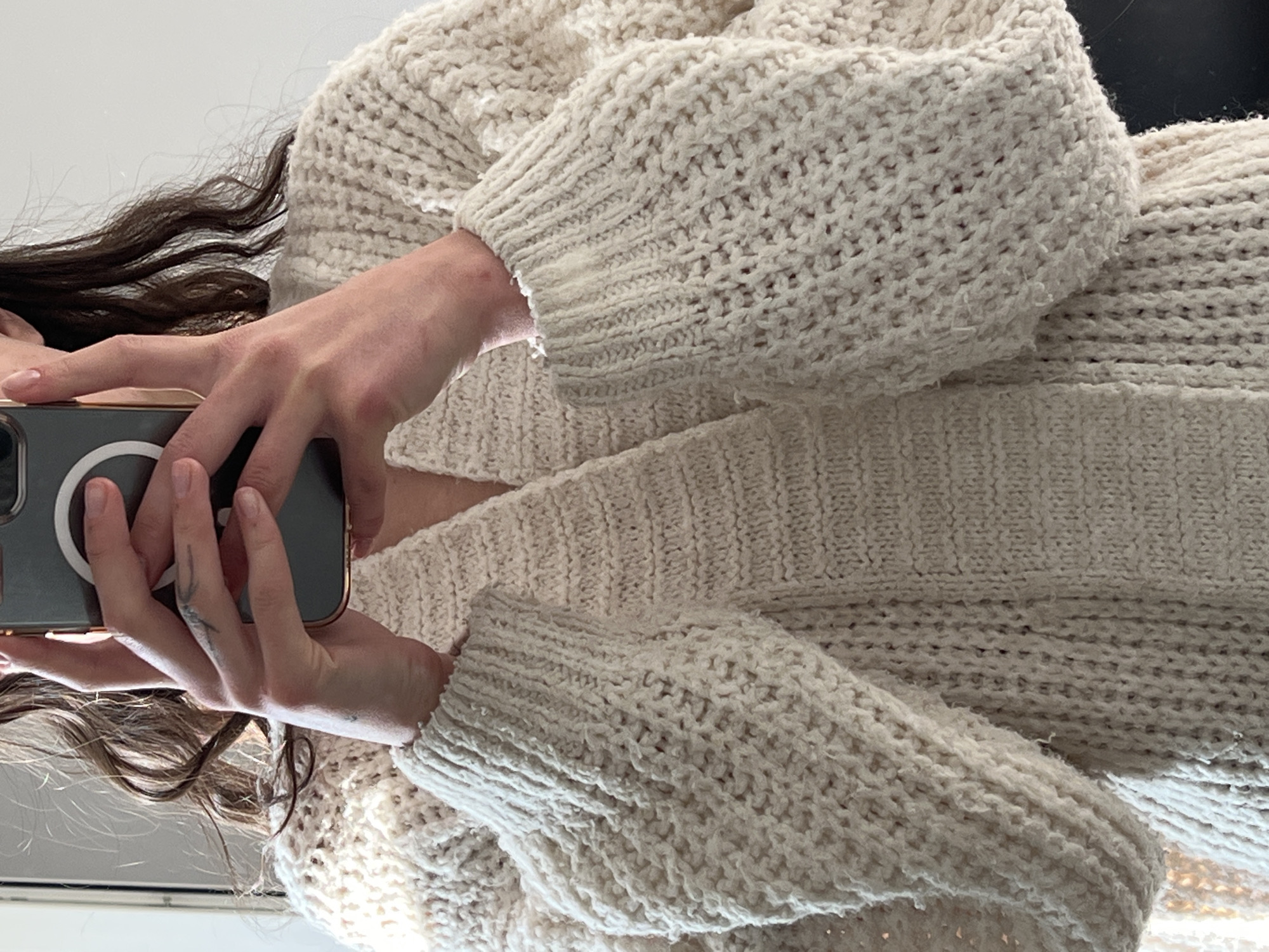 An individual is holding a smartphone, capturing an image of themselves wearing a chunky knit cardigan. The cardigan appears to be cream or off-white in color. The individual has long, dark hair visible near their face and hands. The setting is undefined; there are no visual indicators of the city of Ternopil, Ukraine, or any other specific location.