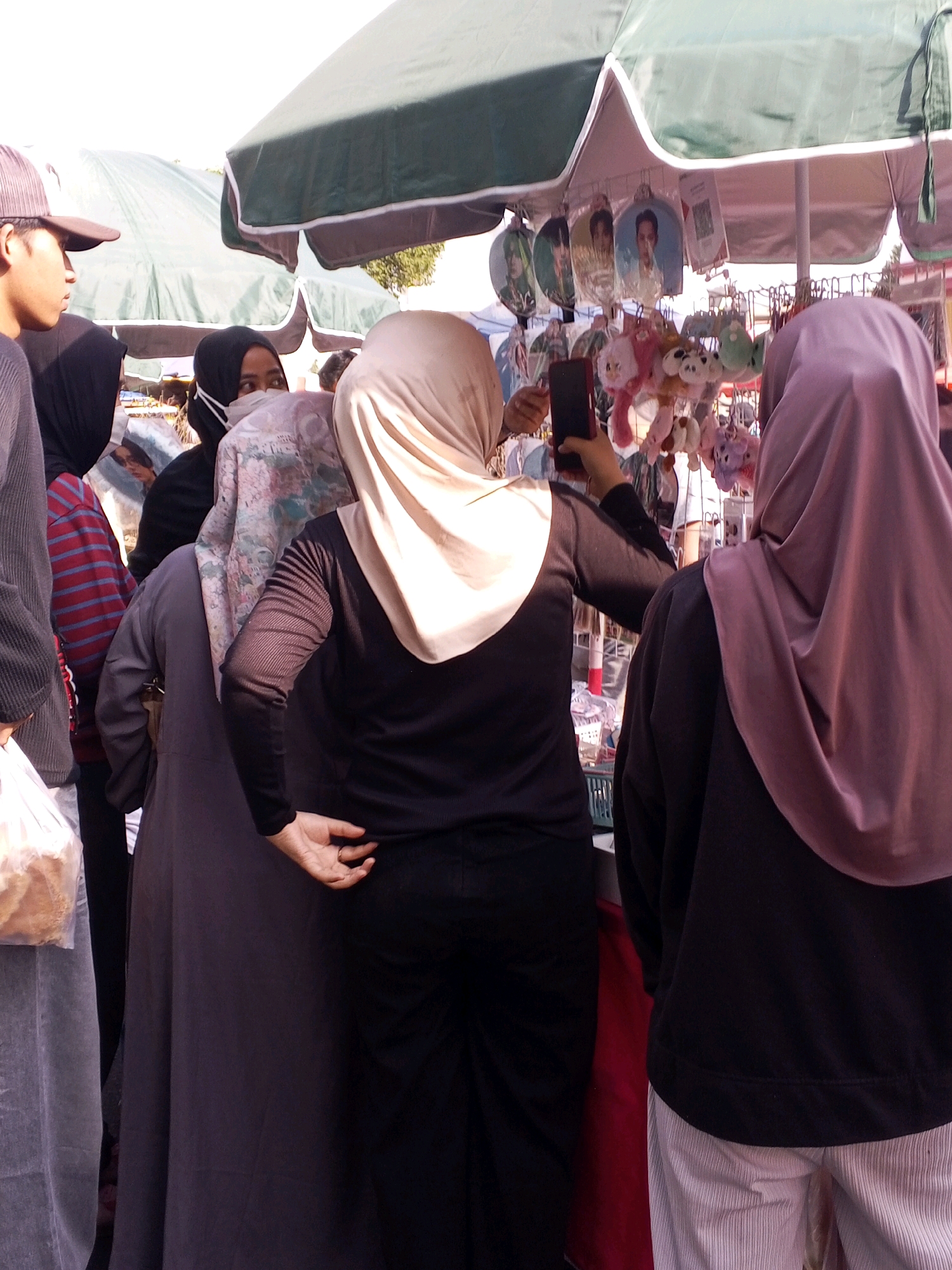 The image depicts a street-level view of a market stall in Serang, Indonesia, likely on a sunny day given the bright lighting and shadows. Several people are visible, predominantly women wearing hijabs, suggesting a cultural context.
In the foreground, a person on the right wears a lilac-colored hijab and a dark long-sleeved shirt, their back partially turned to the viewer. Their lower body is covered by light-colored, vertically striped pants. To the left of this individual, another person wearing a beige hijab and a black long-sleeved shirt is seen from the back, holding a dark smartphone. Their left hand is partially visible, resting on their hip, with a ring on one finger. They are wearing black pants.
Behind these two individuals, a person in a dark grey outer garment and a patterned hijab is visible from the side. To their left, another person is wearing a black hijab. Partially visible to the far left is a person wearing a dark grey long-sleeved garment and a brown baseball cap, with their profile facing left. This person is holding a white plastic bag containing what appears to be bread.
The market stall itself features an umbrella providing shade. Hanging from the stall are various items, including small stuffed animals and round decorative items with images of people on them, possibly merchandise related to popular culture. A green grid basket is also visible. In the background, other market stalls and people are indistinctly present, indicating a bustling market environment. The overall scene is one of commerce and everyday life in an Indonesian city.