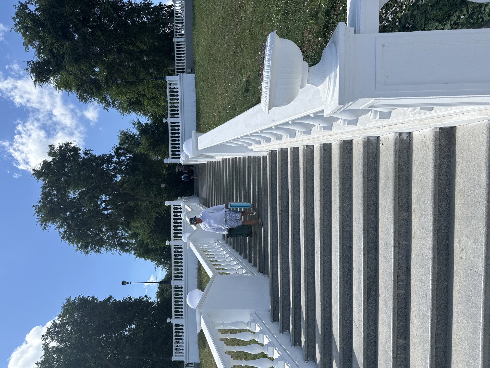 A full, eye-level shot shows a person walking down a wide, ornate, white stone staircase lined with white balustrades and planters. The person, dressed in a white long-sleeved shirt, patterned shorts, a bucket hat, and sandals, carries a green duffel bag and a blue mat. The staircase is made of granite with visible grout lines and has a slight sheen. Behind the staircase, a grassy slope is visible, dotted with small white flowers. In the background, lush green trees frame a bright blue sky with scattered white clouds. A lamppost stands between two trees in the upper left portion of the image. The overall scene depicts a sunny day in an outdoor public space, likely a park or a scenic overlook, possibly within the city of Taman', Russia, given the architectural style of the staircase and the surrounding vegetation.
