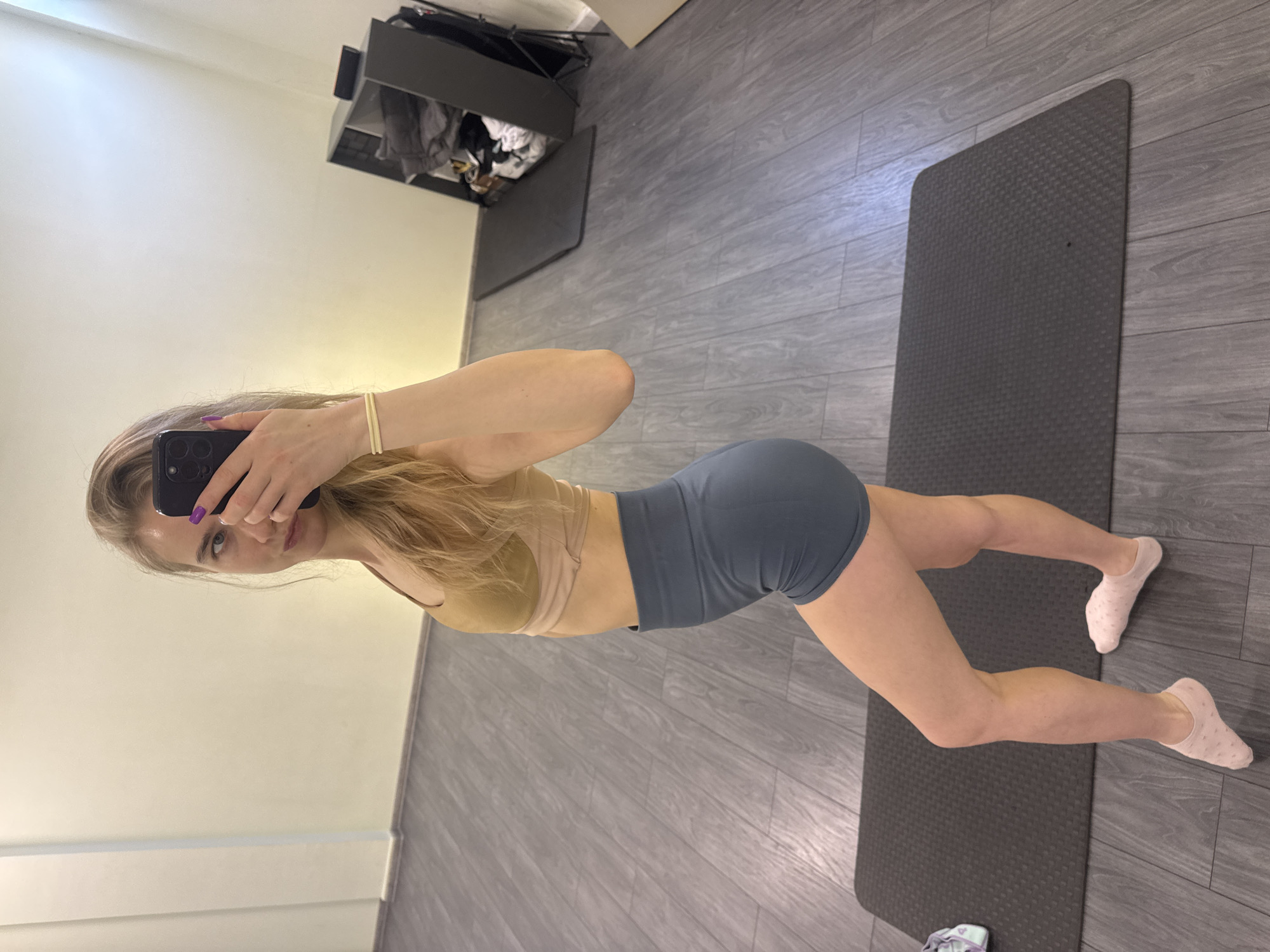 A woman is standing on a grey exercise mat in a room with grey wood-look flooring and a beige wall. She is wearing a tan sports bra and grey exercise shorts and has on light pink socks. She is holding a phone in her left hand and taking a picture of herself. There is a grey storage unit or cabinet in the corner of the room. The location is Unknown, Unknown.