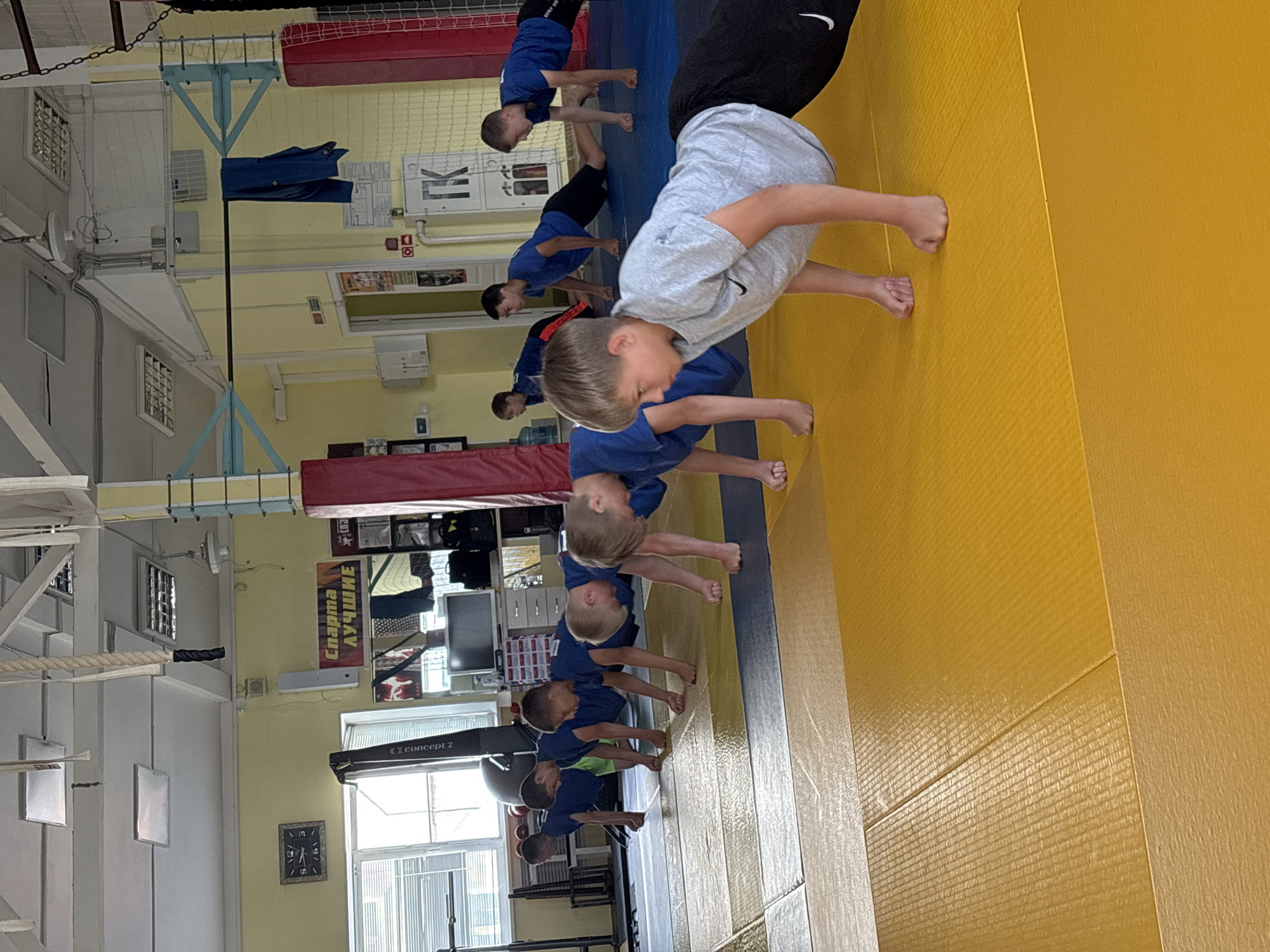 In a gymnasium, likely located in Chaikovsky, Russia, a group of young males are performing push-ups on yellow and blue mats. They are positioned in a line, facing away from the camera, with their hands on the floor and bodies extended. The environment features a yellow-painted wall, a clock on the wall displaying 12:17, a large window with blinds, a rowing machine, and various sports equipment including red and blue mats, and a rope hanging from the ceiling. There are also posters on the wall. The boys are wearing athletic attire.