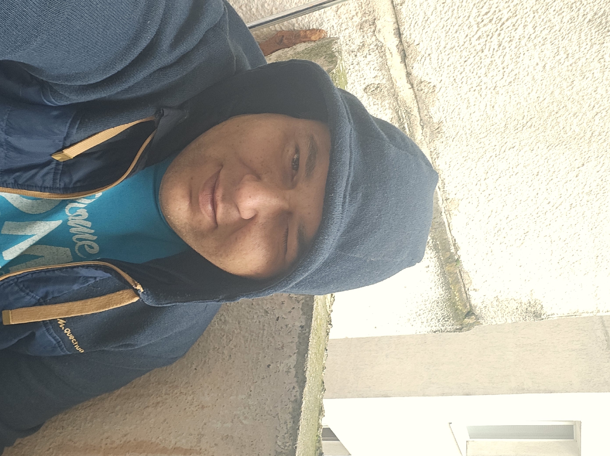 A man wearing a blue hooded jacket and a blue t-shirt is positioned outdoors, possibly near a building. The jacket has tan-colored straps and the text "M. Quechua" is visible on the garment. He is facing the camera with a slight smile, and only one eye is partially visible.
The background consists of a textured wall with variations in color, indicating possible weathering or staining. A segment of a window frame or part of a building is present in the lower right corner. The specific location within Stara Zagora, Bulgaria cannot be determined from the provided visual information.