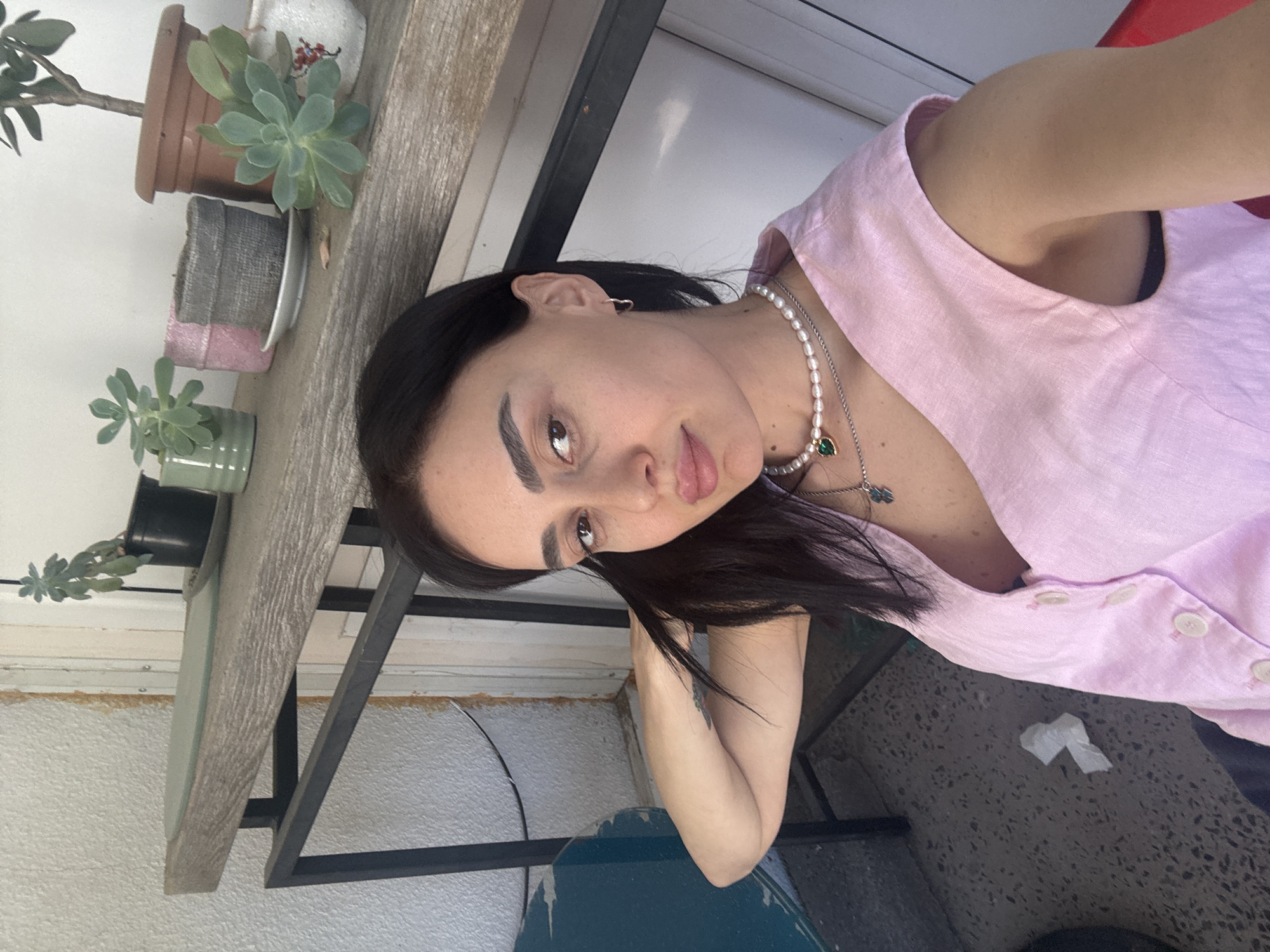 A woman with dark hair, wearing a pink sleeveless button-down shirt and a pearl necklace with a green stone, is posing for a photograph. The setting appears to be a balcony or outdoor seating area with potted plants along a weathered wooden surface. A gray tiled floor is partially visible. Given the provided information, this image was likely taken in Tbilisi, Georgia.