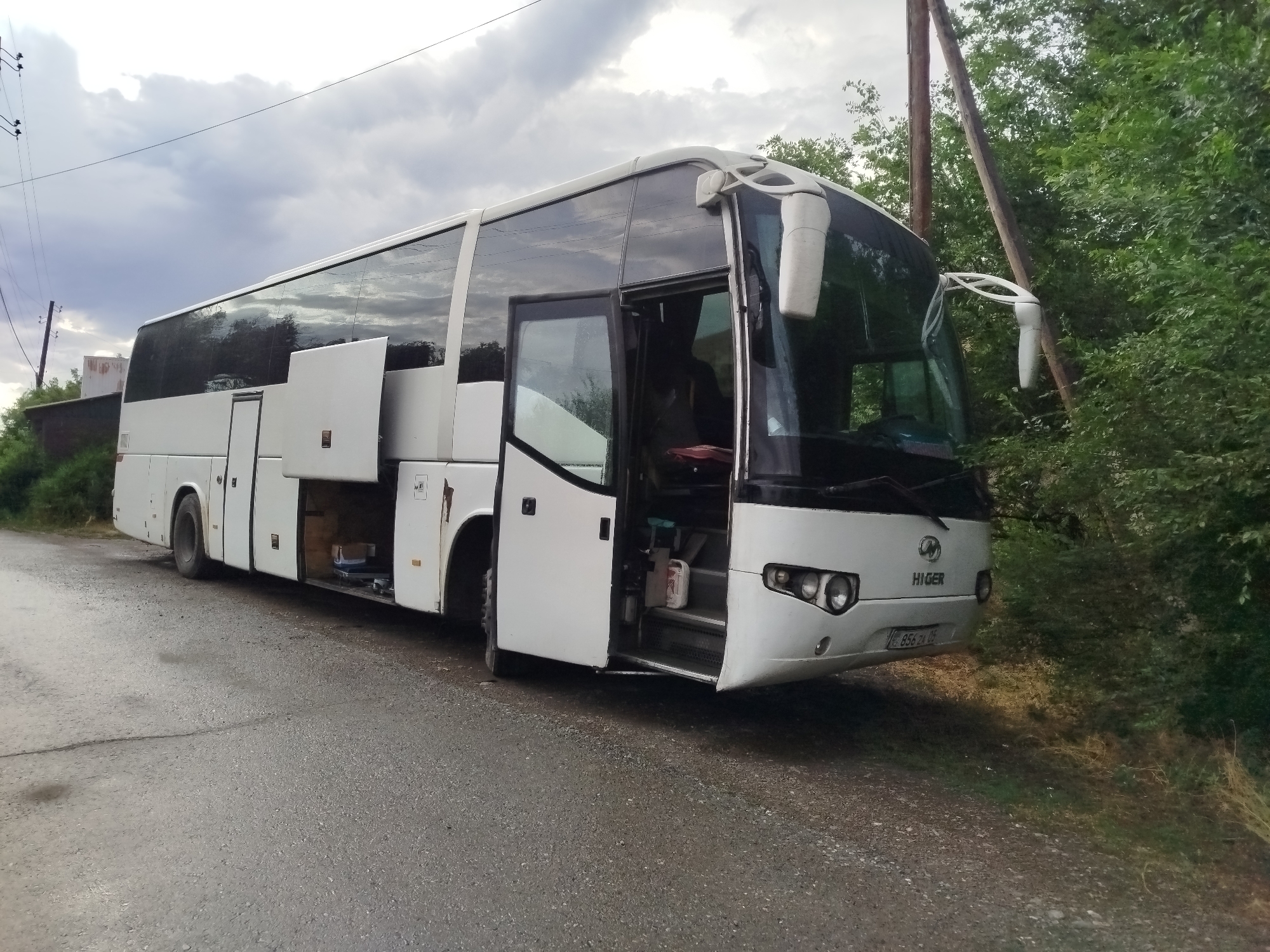 A white Higer bus is parked on the side of a paved road in Kabanbay, Kazakhstan. The bus's front passenger door is open, revealing the steps leading into the cabin. Several storage compartments along the side of the bus are also open, exposing their contents. The bus is surrounded by lush green trees and vegetation, with power lines visible in the background. The sky is overcast with dramatic clouds. The license plate of the bus is visible and reads "856 ZX 05".