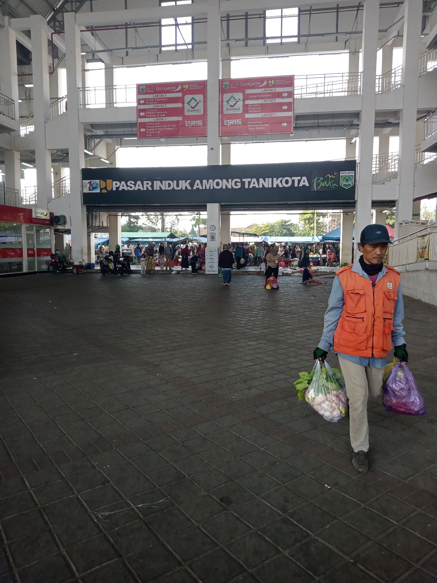 The image captures an outdoor scene at the Pasar Induk Among Tani in Batu, Indonesia. The main subject, a man wearing an orange vest and a dark baseball cap, walks from right to left, carrying two plastic bags filled with groceries. He is dressed in a light blue long-sleeved shirt and beige pants. Behind him, the market is bustling with activity. People can be seen browsing stalls, and there are motorcycles parked in the foreground. A large sign above the market entrance reads "PASAR INDUK AMONG TANI KOTA BATU" with the city's emblem. The architecture suggests a covered market with large pillars and an elevated walkway. The ground is paved with dark, rectangular tiles. The lighting indicates it is daytime.