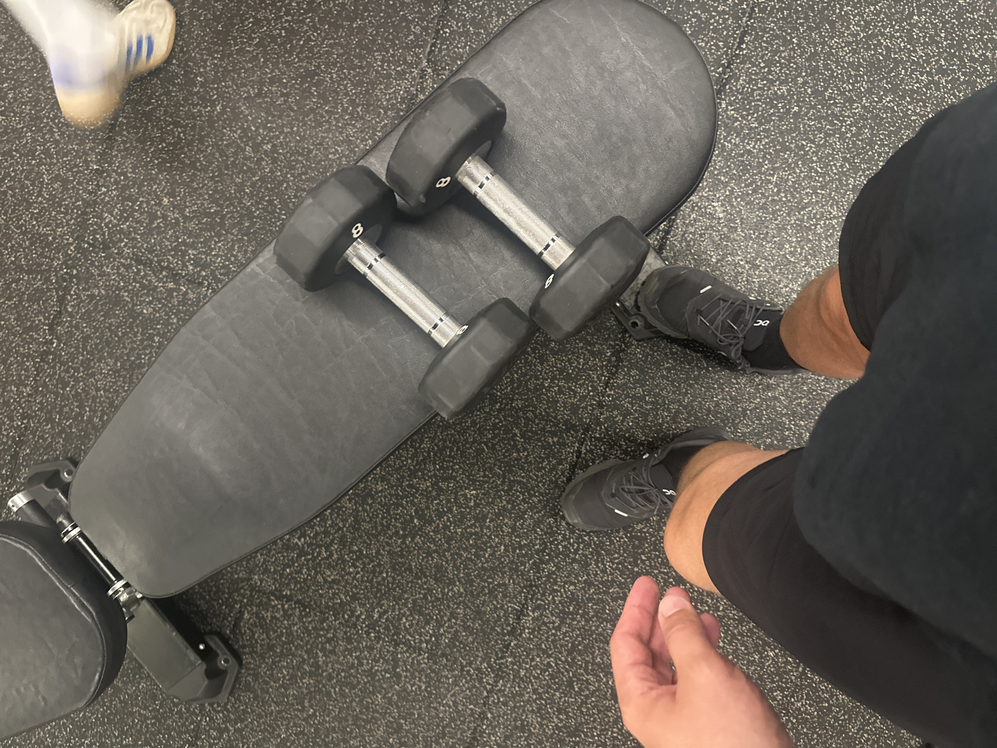 A first-person, high-angle shot displays a section of a gym setting. In the center is a black adjustable weight bench. Two 8 lb dumbbells rest on the bench. The floor is a speckled gray rubber material. The legs of the person taking the photo are partially visible on the right. They wear black shorts and black running shoes. A foot of another person wearing a white and blue sneaker is visible in the top left corner. The location of the image is not identifiable.