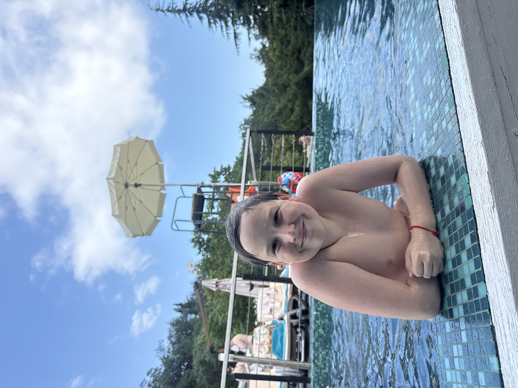 A young boy with wet hair and a smile is leaning on the edge of a swimming pool. He is shirtless and wearing a red string bracelet on his left wrist. The pool has a tiled edge with blue and green tiles. In the background, there is a lifeguard stand, umbrellas, trees, and a blue sky with clouds. The scene appears to be set in a sunny outdoor location, likely a resort or a public pool, given the presence of other people and amenities. The overall mood is cheerful and relaxed, suggesting a day of swimming and enjoyment. The location context provided indicates the setting is Yalta, Ukraine.