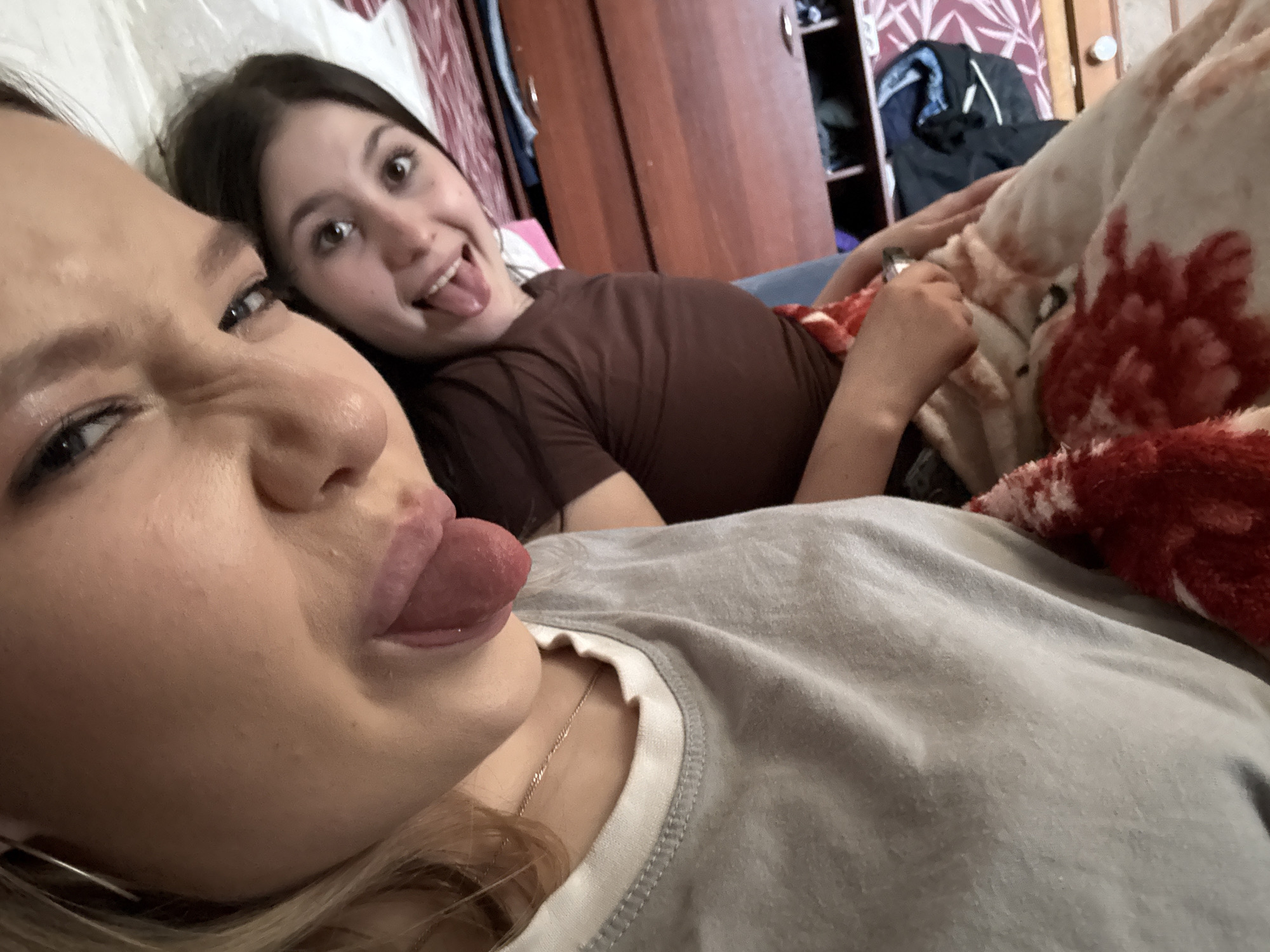 Two young women are posing in a selfie. The woman in the foreground is fair-skinned with blonde hair, and she is sticking out her tongue. She wears a light grey T-shirt with white trim. The woman in the background has dark brown hair and is wearing a brown shirt and is sticking out her tongue. They appear to be in a bedroom. The location is Фоки, Russia. A patterned blanket and a closed wooden wardrobe are visible in the background.