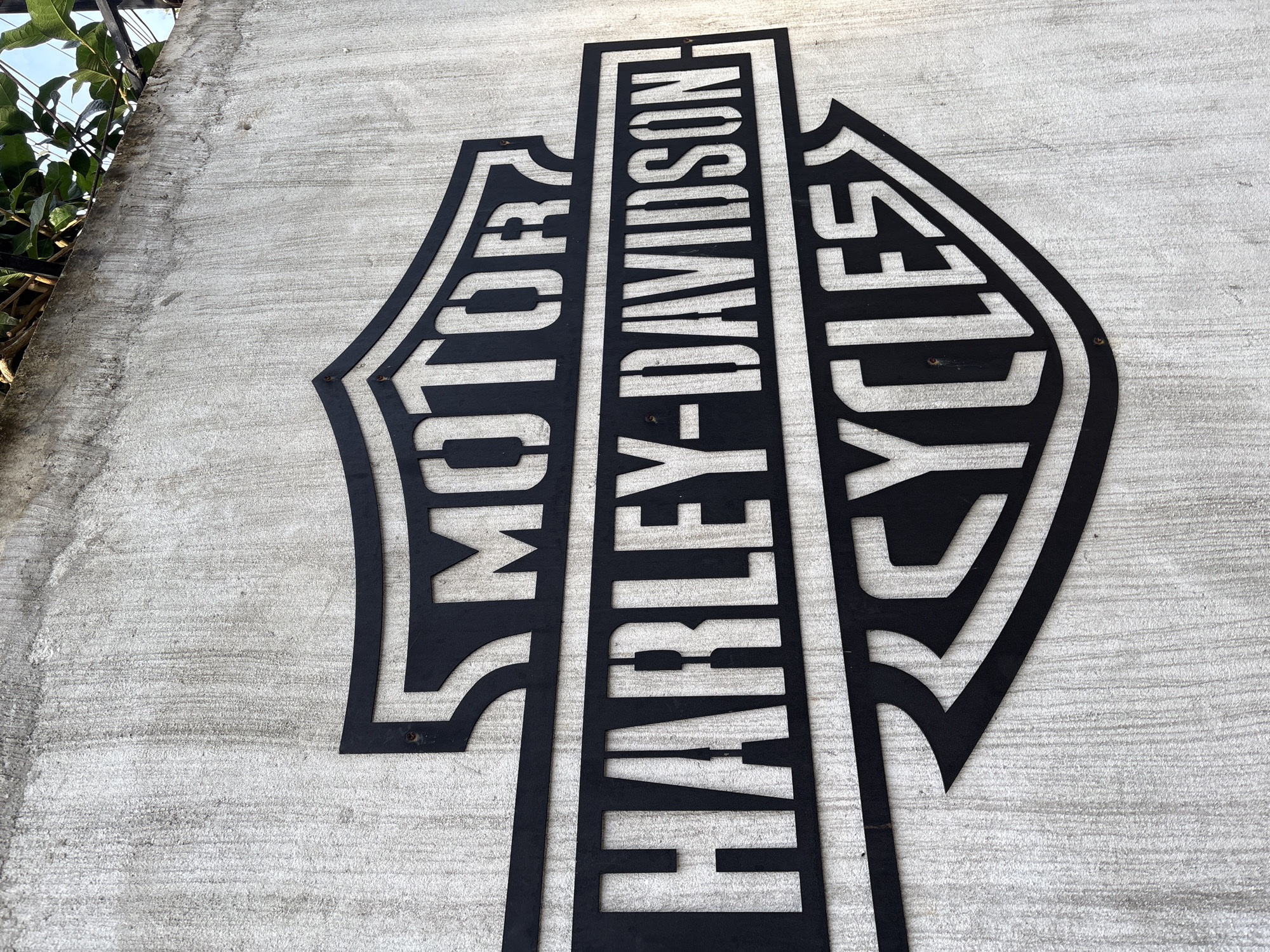 A close-up, low-angle shot shows a black metal Harley-Davidson Cycles logo mounted on a rough, textured concrete wall. The logo is a shield shape with "MOTOR" in the upper portion, "HARLEY-DAVIDSON" in the middle horizontal bar, and "CYCLES" in the lower part within a wing-like design. The concrete wall appears weathered, with vertical streaks and some discoloration. The top edge of the wall is visible, with green foliage and a black metal fence encroaching from above, suggesting an outdoor setting. The image is well-lit, with shadows cast by the metal logo onto the wall. This scene is located in Karon, Thailand.