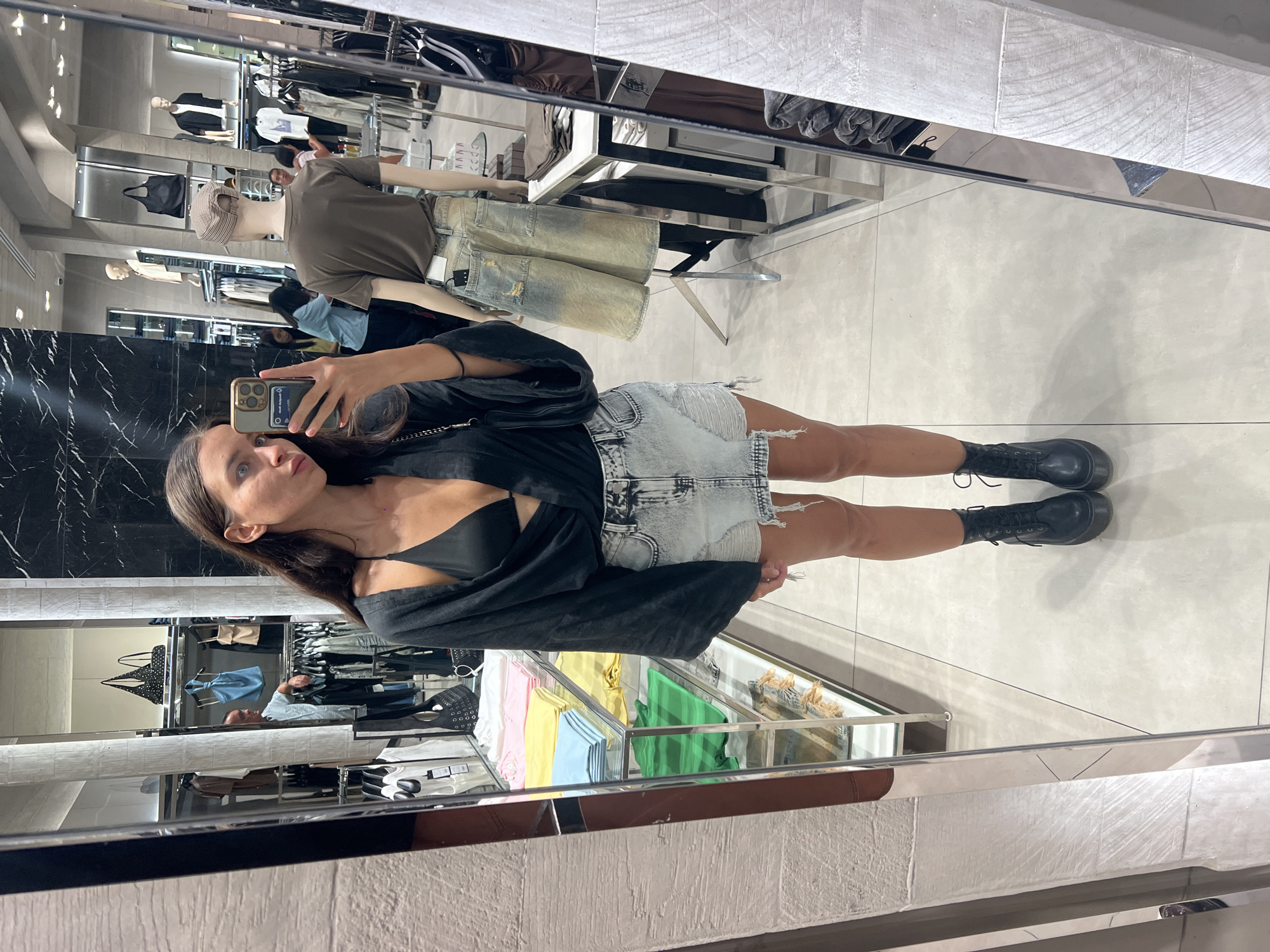 The image shows a woman taking a mirror selfie in a retail environment. The location appears to be a clothing store, with mannequins displaying apparel in the background. The woman is wearing a black top, a light-colored denim skirt, and black boots. She is holding a smartphone up to capture the image. The mirror reflects the store's interior, including racks of clothing and other customers. The scene is well-lit and orderly, typical of a retail setting.