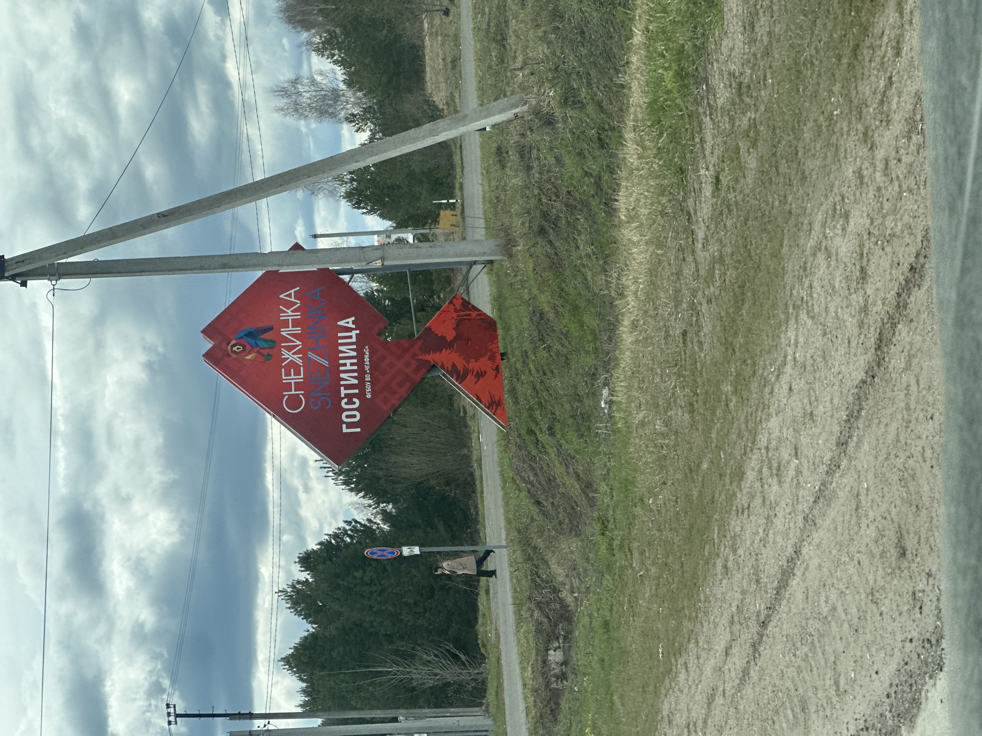 The image, taken in Priamsky, Russia, shows an outdoor scene featuring a large red advertising sign, utility poles, a natural landscape, and a person.
In the foreground, a prominent red, diamond-shaped sign is supported by two concrete utility poles forming an inverted V-shape. The sign displays text in Cyrillic. The primary text reads "СНЕЖИНКА" (Snezhinka), meaning "snowflake," and below it, "ГОСТИНИЦА" (Gostinitsa), meaning "hotel." There is also smaller text at the bottom right, "ФГБОУ ВО «ЧГПУ им. И.Я. Яковлева»", which refers to the "Chuvash State Pedagogical University named after I. Ya. Yakovlev." The sign features a stylized, colorful logo with abstract shapes, possibly representing a snowflake or a bird.
To the right of the sign, there is a grassy area with patches of bare dirt, and a visible dirt path or track runs parallel to the main road. Further back, a line of trees, some deciduous and bare, others evergreen, marks the horizon. The sky is overcast with a mix of light and darker grey clouds, indicating a cloudy daytime setting.
A single person is visible walking on the dirt path, further down from the main sign. They are wearing a light-colored coat or jacket and dark trousers, appearing to walk in the direction of the sign. Several power lines stretch across the frame, connected to other utility poles in the distance. The overall scene suggests a rural or semi-rural roadside in temperate weather.