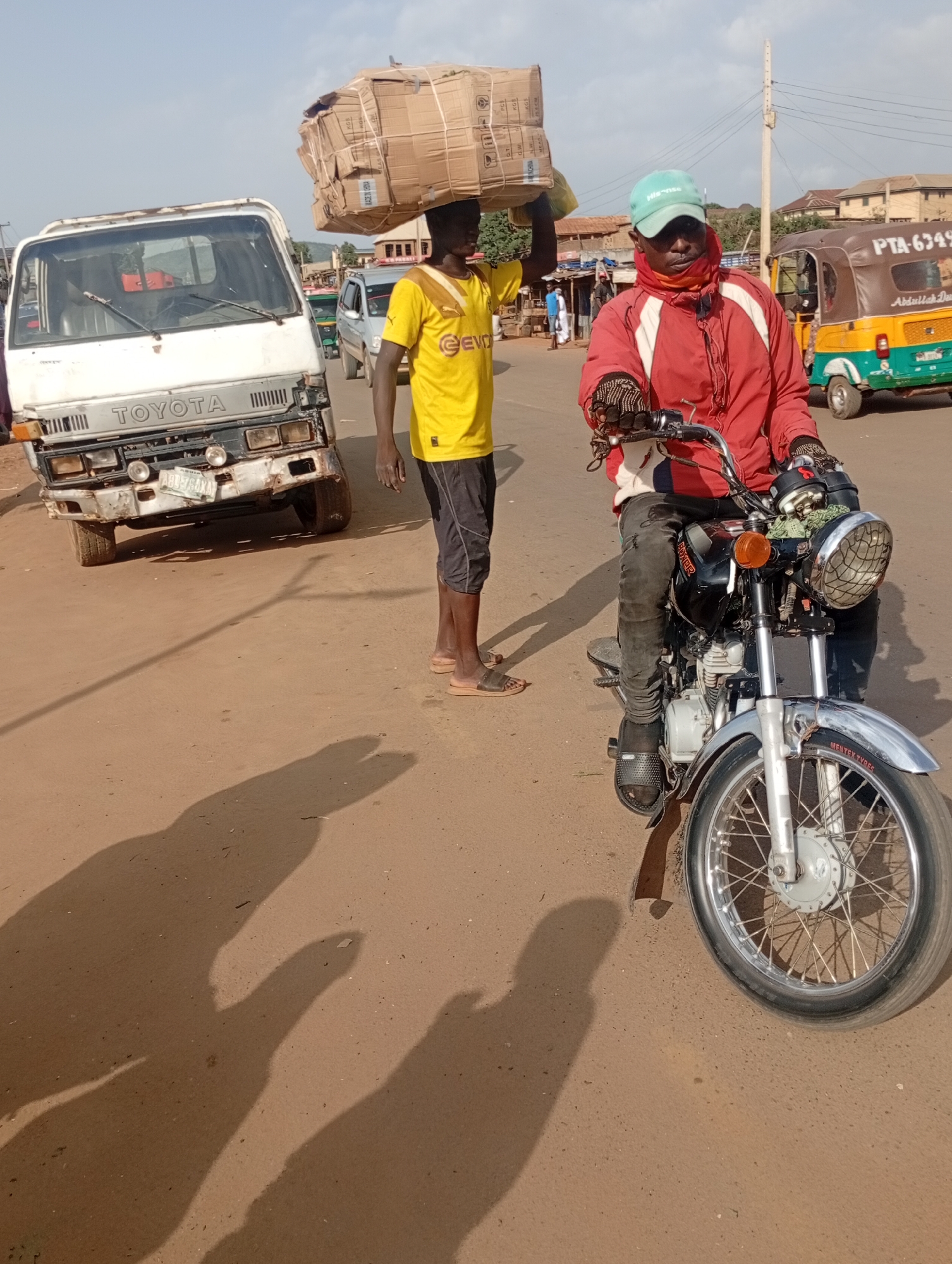 In Rafin Par, Nigeria, a man wearing a yellow t-shirt and dark shorts carries a large cardboard box on his head. He stands on a dusty road next to a white Toyota truck. To his right, another man rides a black motorcycle. He wears a red jacket, dark pants, and a light blue cap. The motorcycle has a chrome fender and a round headlight. In the background, a multi-colored auto-rickshaw is parked, with text partially visible. Buildings and trees line the street, suggesting an urban or semi-urban environment. The sun is high, casting long shadows on the road, indicating it is likely daytime. The overall scene depicts everyday life and transportation in Nigeria.