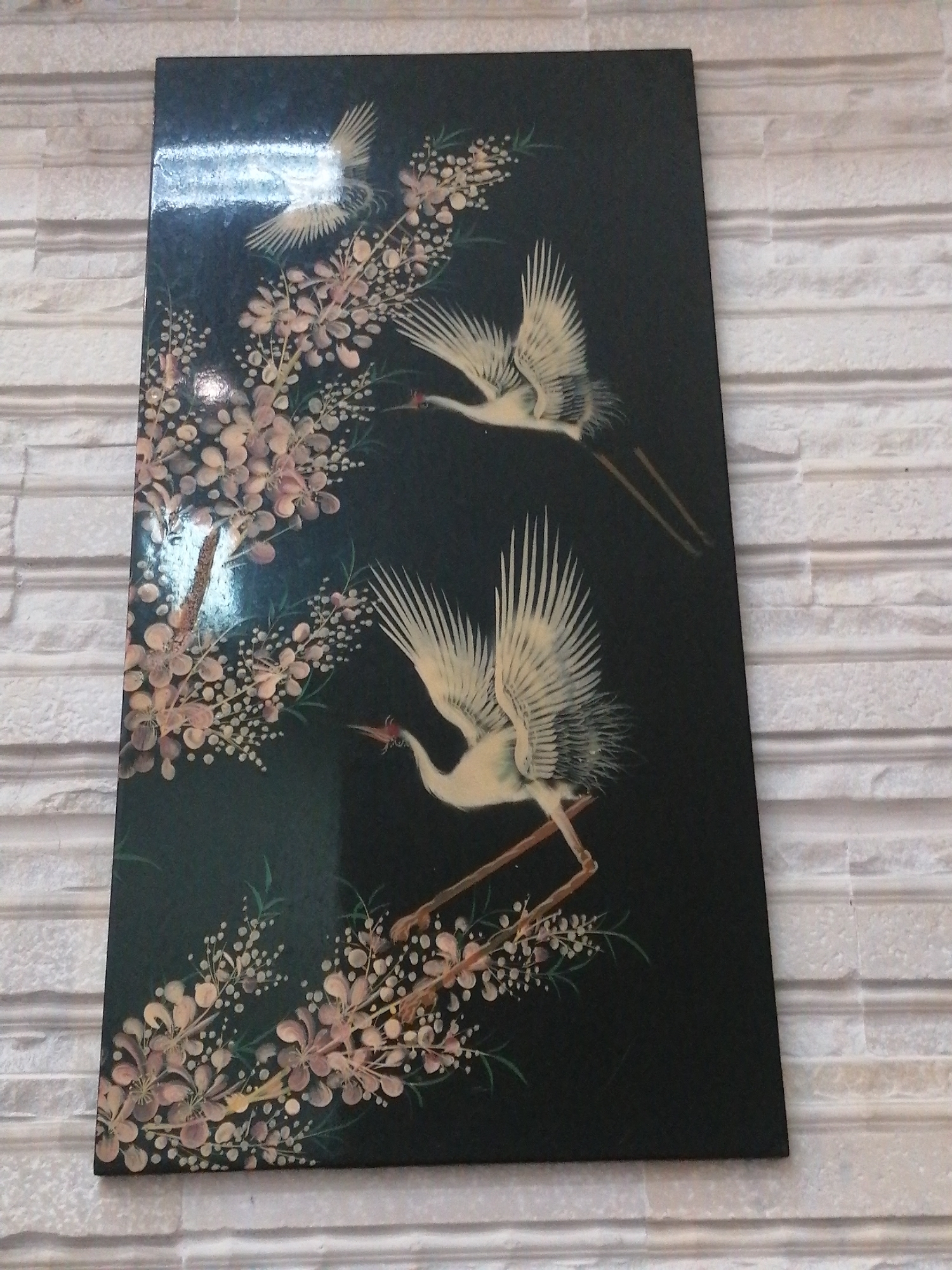 A rectangular black lacquered panel, likely a decorative screen or wall art, depicts cranes and flowering branches. The panel is leaning against a white brick wall. In the foreground, a large crane with white feathers and a red beak is in flight, its wings spread wide, with its legs extended. Behind it and slightly higher, another crane in a similar pose is visible, though less detailed. In the upper left corner, a third, smaller crane is depicted in flight. Delicate branches with small, pale pink and white blossoms and tiny white buds are interspersed throughout the scene, with some branches reaching across the panel. The background is a solid, glossy black. The image is taken from a slightly elevated, oblique angle.