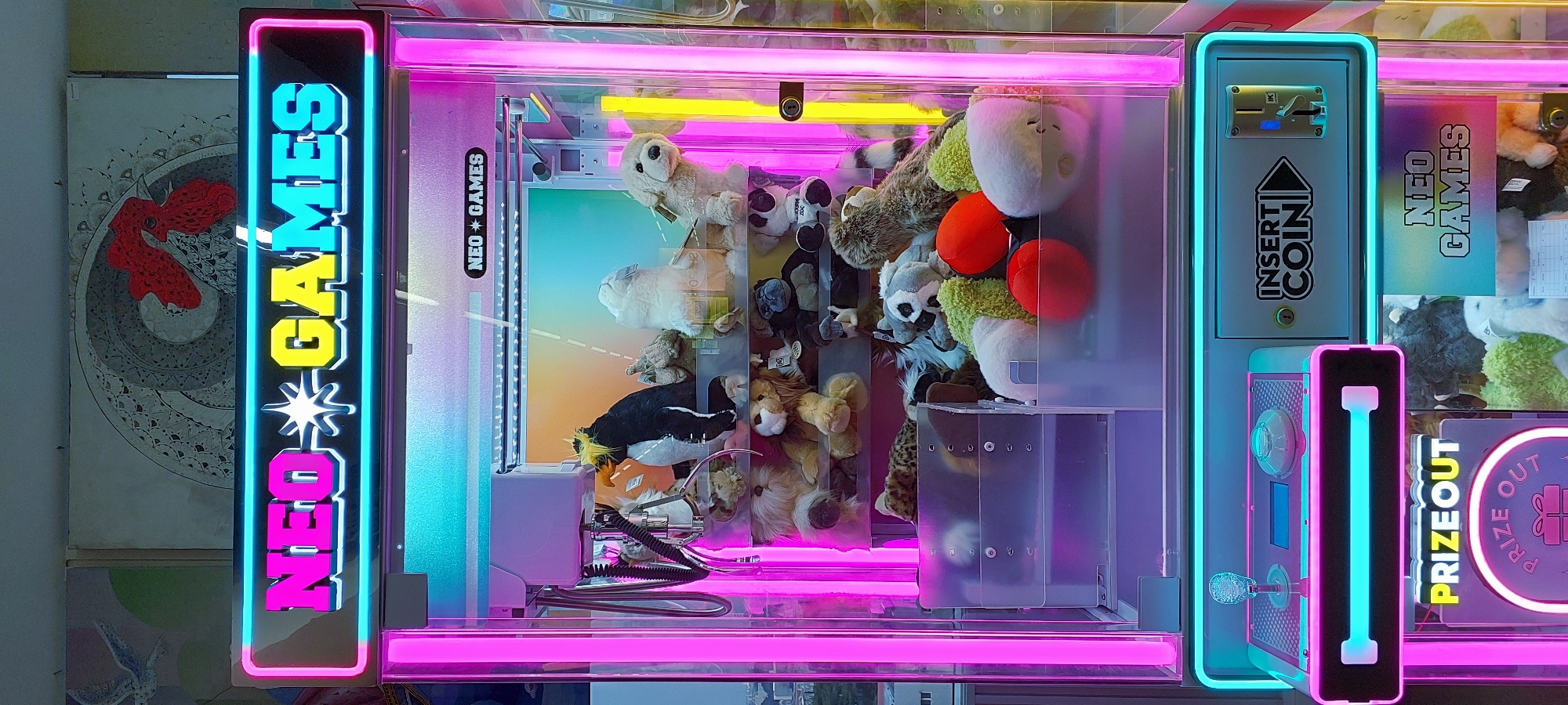The image shows a vertical, well-lit claw machine filled with plush toys, located in Curug Sangereng, Indonesia. The machine features bright neon pink and cyan lighting, with "NEO GAMES" displayed prominently in illuminated letters at the top. Inside, an assortment of stuffed animals, including a penguin, dogs, a lion, a monkey, and various other characters, are arranged on shelves. A claw mechanism is visible above the toys. The control panel at the bottom includes a joystick, buttons, and coin slots labeled "INSERT COIN." Below the control panel, a "PRIZE OUT" chute is also visible. The background suggests an indoor setting, possibly an arcade or entertainment center, with a blurred wall or display behind the machine. The overall scene is a brightly lit, commercial entertainment environment.