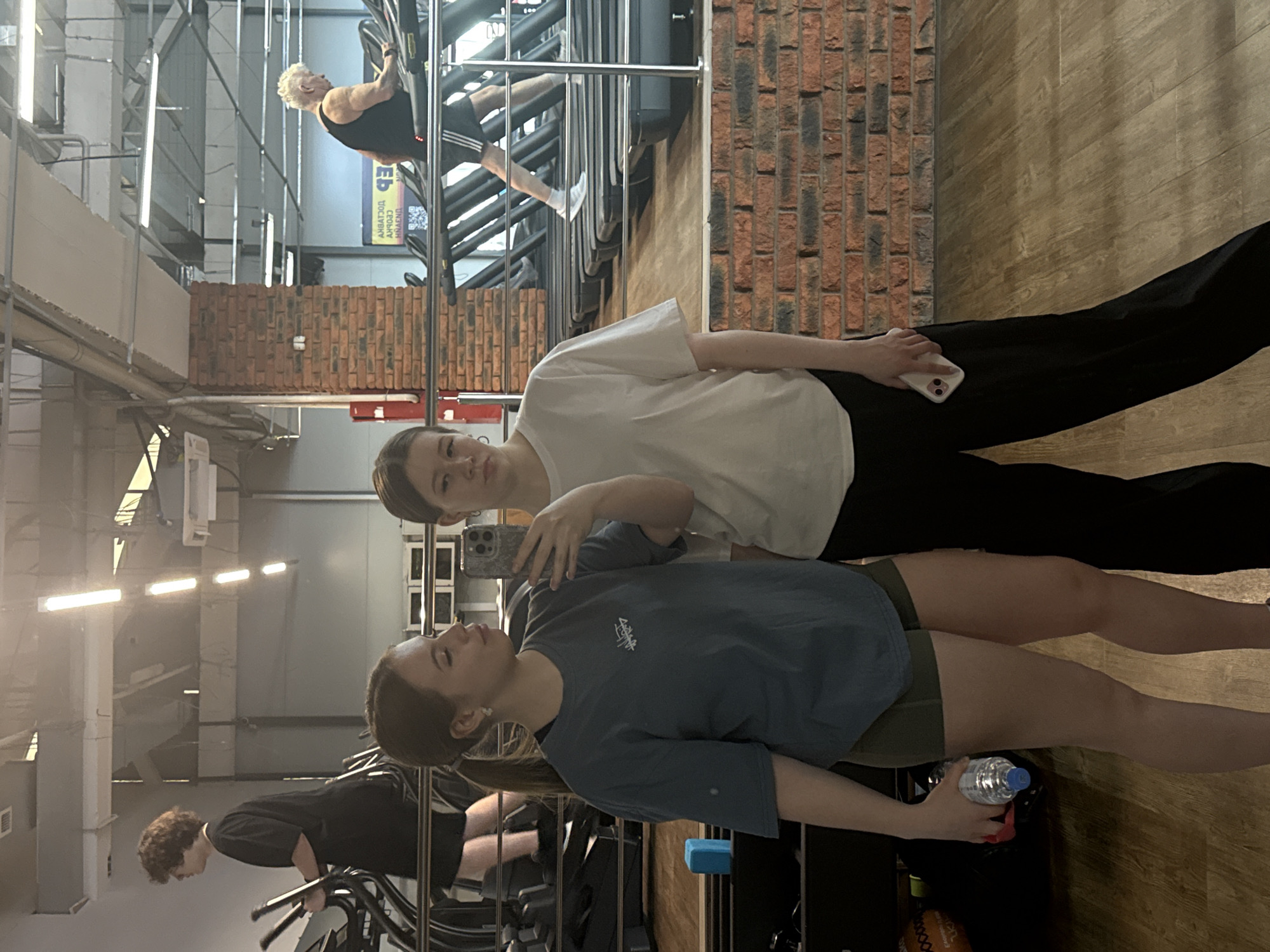 Two young women are captured in a mirror selfie within a modern gym setting. In the foreground, the woman on the left wears a blue t-shirt with a white logo and olive green shorts, holding a plastic water bottle with a blue cap. Beside her, the woman on the right wears a white t-shirt and black pants, holding a light-colored smartphone in her left hand, which she is using to take the picture.
The background, a reflection in the mirror, reveals various gym activities and architectural details. An older individual with short white hair, dressed in a black tank top and black shorts with white stripes, is actively engaged on a climbing-like exercise machine. Further back, parts of other gym-goers are visible, including the lower legs and feet of a person in white socks and dark shorts, presumably on a treadmill, and a young man with curly brown hair in a black t-shirt, operating another piece of equipment.
The gym features an industrial design with exposed ventilation ducts, bright linear light fixtures on the ceiling, metal structural elements, and accent walls made of red brick. The floor is covered with wooden planks. Visible on a sign reflected near the climbing machine is partially obscured and mirrored Cyrillic text, which, when corrected, appears to read "СПОРТ" (SPORT) and likely "СКИДКИ" (DISCOUNTS), suggesting promotional information. The scene depicts a lively atmosphere in a well-lit indoor fitness center.