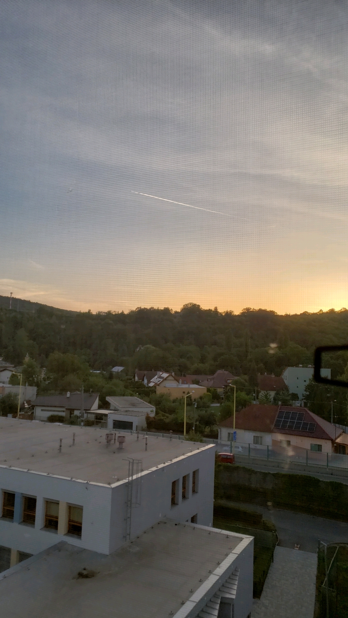 This is a vertical, eye-level shot taken from a window covered with a screen. It captures a suburban landscape at sunset. The sky is a gradient of pale blue at the top, transitioning to a soft orange glow near the horizon, with a thin contrail visible high up, indicating an airplane passed overhead. Below the sky, a dense, tree-covered hillside forms the backdrop. A small settlement of houses with red-tiled roofs is nestled at the base of the hill. In the foreground, there is a multi-story building with a flat roof, showing a fire escape ladder on its side. Another flat-roofed structure is partially visible in the lower part of the frame. A small red car is parked on the road in the middle ground. The overall impression is of a quiet, residential area as evening approaches.