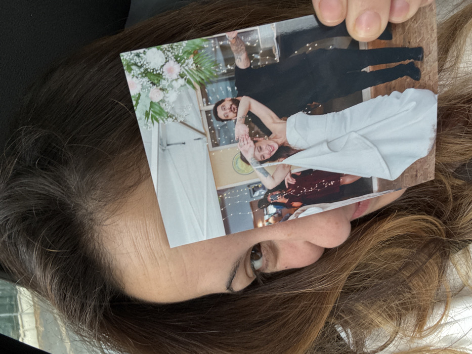 An individual with brown hair, partially visible on the right side of the frame, holds a glossy color photograph in a close-up, selfie-style shot taken in Arcueil, France. The person's right eye, nose, and part of their mouth are visible on the left side, as their left hand grips the bottom right corner of the photograph. The background behind the individual is dark and out of focus.
The photograph depicts a celebratory indoor event featuring two primary subjects. A female, centrally positioned, wears a white strapless gown with a draped skirt. She has dark hair, visible tattoos on her left forearm, and is smiling while raising her clasped hands above her head. To her right, a bearded male with dark hair is dressed in a black t-shirt and black pants, displaying tattoos on his arms. He smiles broadly with his arms extended outward, palms up. In the background of the photograph, a third person in a dark red dress is visible to the left of the female in white, appearing to hold a camera or phone. The depicted environment includes white canopy structures, string lights, and floral arrangements of pink and white flowers with green foliage.
