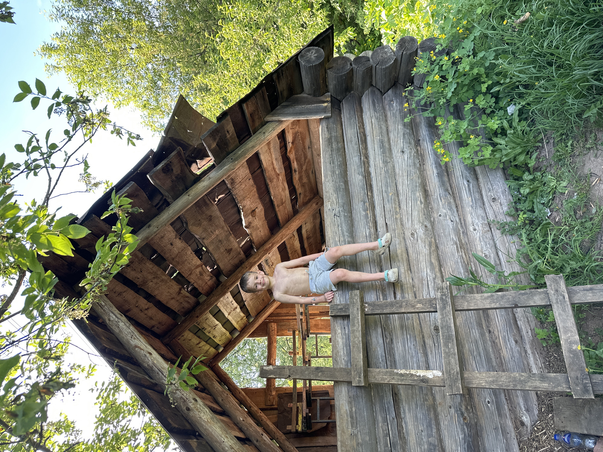 A child lies on the wooden platform of a dilapidated structure in Alnyash, Russia. The structure, built from logs and planks, appears to be some kind of shed or small cabin with a partial roof. The child is shirtless, wearing shorts and sandals. The surrounding environment includes trees and vegetation. The image is taken from a low angle, looking up at the structure.