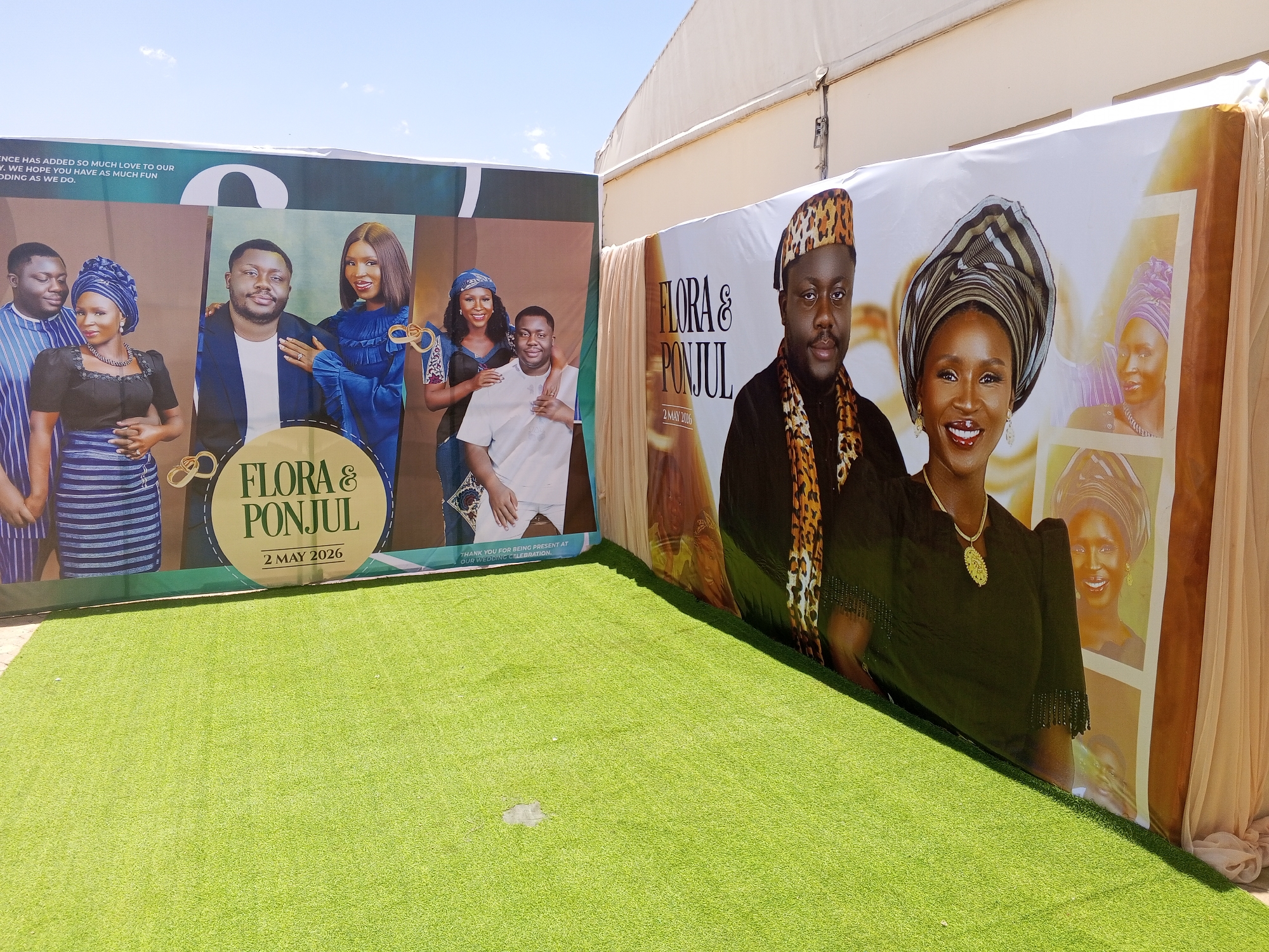 The image shows two printed banners, side-by-side, set up outdoors on a green, artificial turf surface.
The banner on the left features multiple photographs of couples. The banner's text reads "FLORA & PONJUL", and "2 MAY 2026".
The banner on the right features a prominent photograph of a man and woman, with smaller inset photos of the woman. The text reads "FLORA & PONJUL" and "2 MAY 2006".
The environment appears to be a courtyard or event space, based on the turf and the glimpses of a building in the background. The scene is well-lit, suggesting an outdoor setting during daylight hours. It is likely a wedding backdrop given the couple photos. There is no indication this photograph was taken in Jos, Nigeria.
