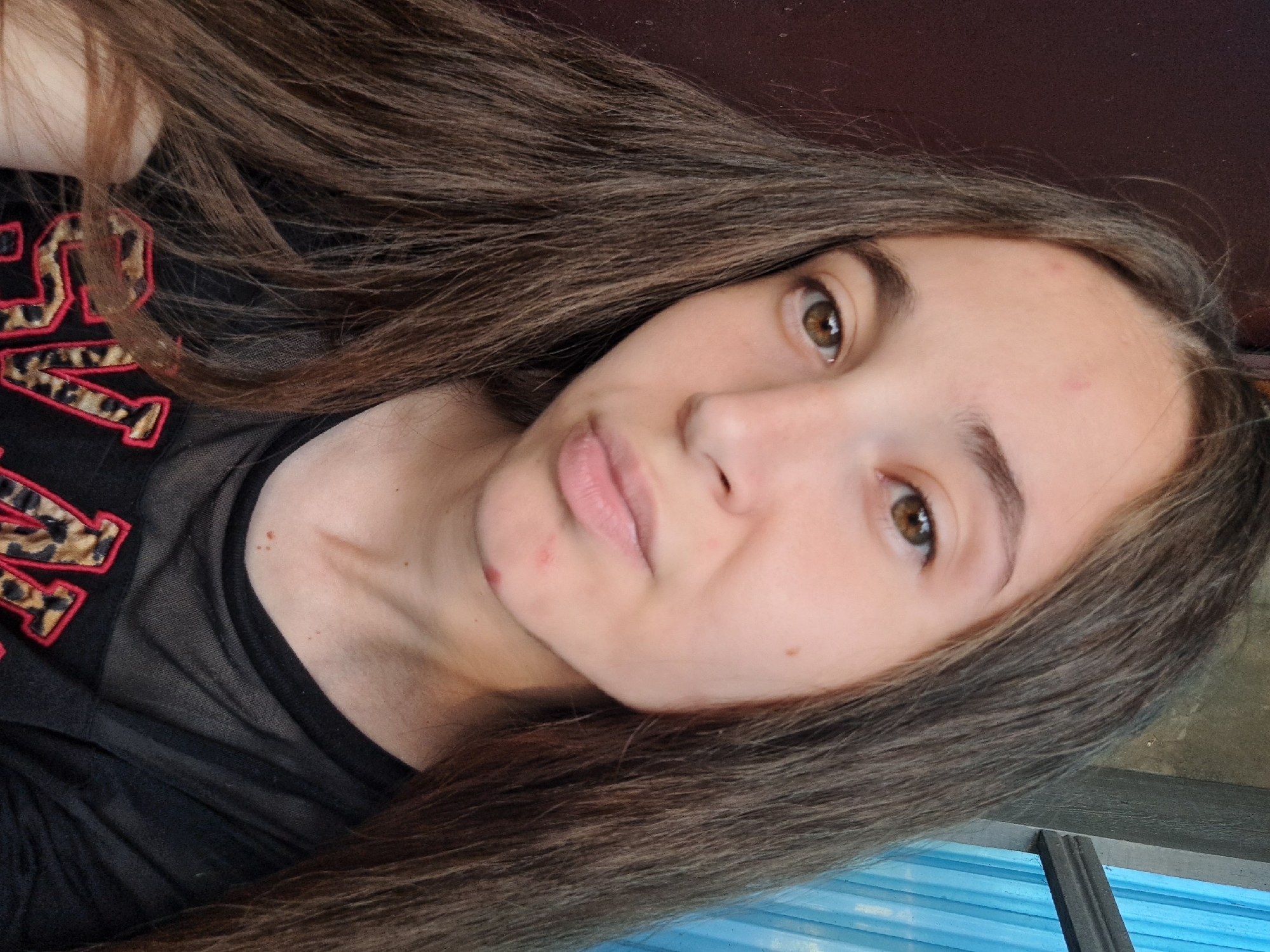 The image is a close-up, front-facing selfie of a young person with long, brown hair. Their skin has a few blemishes, including a red pimple on their forehead and two on their chin. They are wearing a black t-shirt with a sheer neckline and an animal print design with red trim. The background is blurred, with a dark brown wall on the left and a blue curtain and gray wooden structure on the right, suggesting an indoor setting. No specific location in Vypasne, Ukraine is identifiable from the visual information provided.