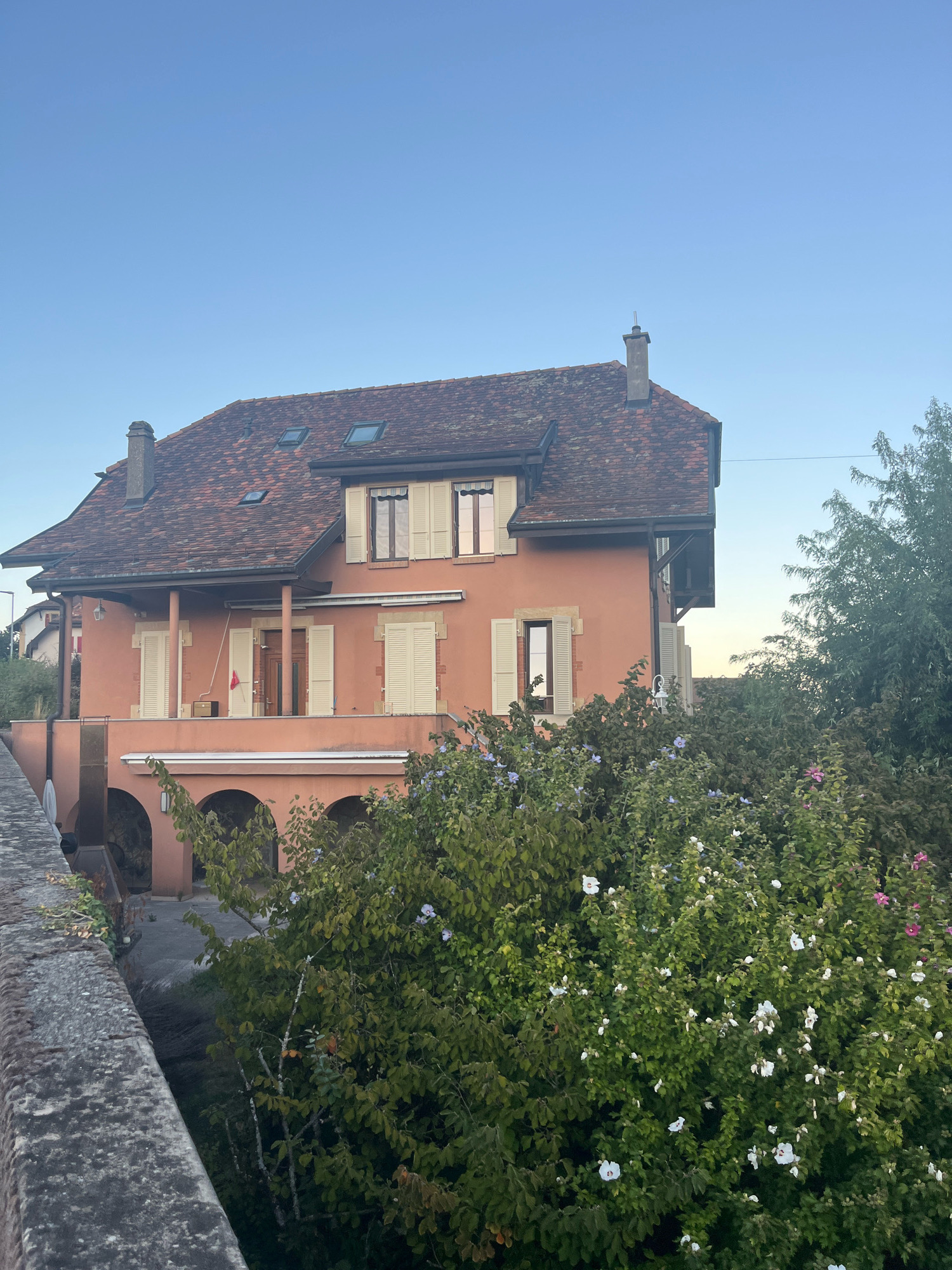 The image depicts a multi-story salmon-colored house with a red-tiled roof and several dormer windows. The house has a covered balcony supported by columns and arches on the lower level. Green foliage with scattered white and pink flowers surrounds the house. A stone wall is visible in the foreground on the left. The scene appears to be set in a residential area with clear blue sky overhead. Based on the context of Féchy, Switzerland, this is likely a private residence in the town, possibly with views of Lake Geneva or the surrounding countryside.