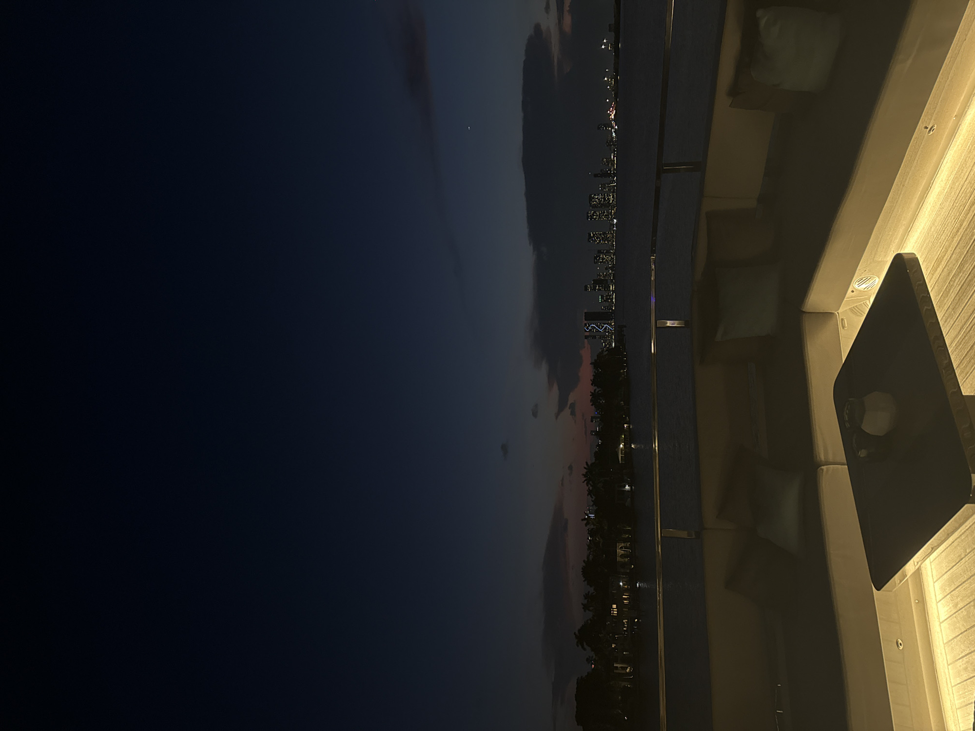 Here is a concise description of the image:
The image depicts a view of Miami Beach, Florida, United States, at night. From what appears to be a rooftop or high vantage point, the city skyline is visible along the horizon. Along the right side of the frame is a portion of a modern outdoor seating area with cushioned benches, and a dark-colored table. The sky transitions from a deep blue on the left to a more illuminated gradient with hints of pink and orange towards the horizon. The city lights create a sparkling effect.