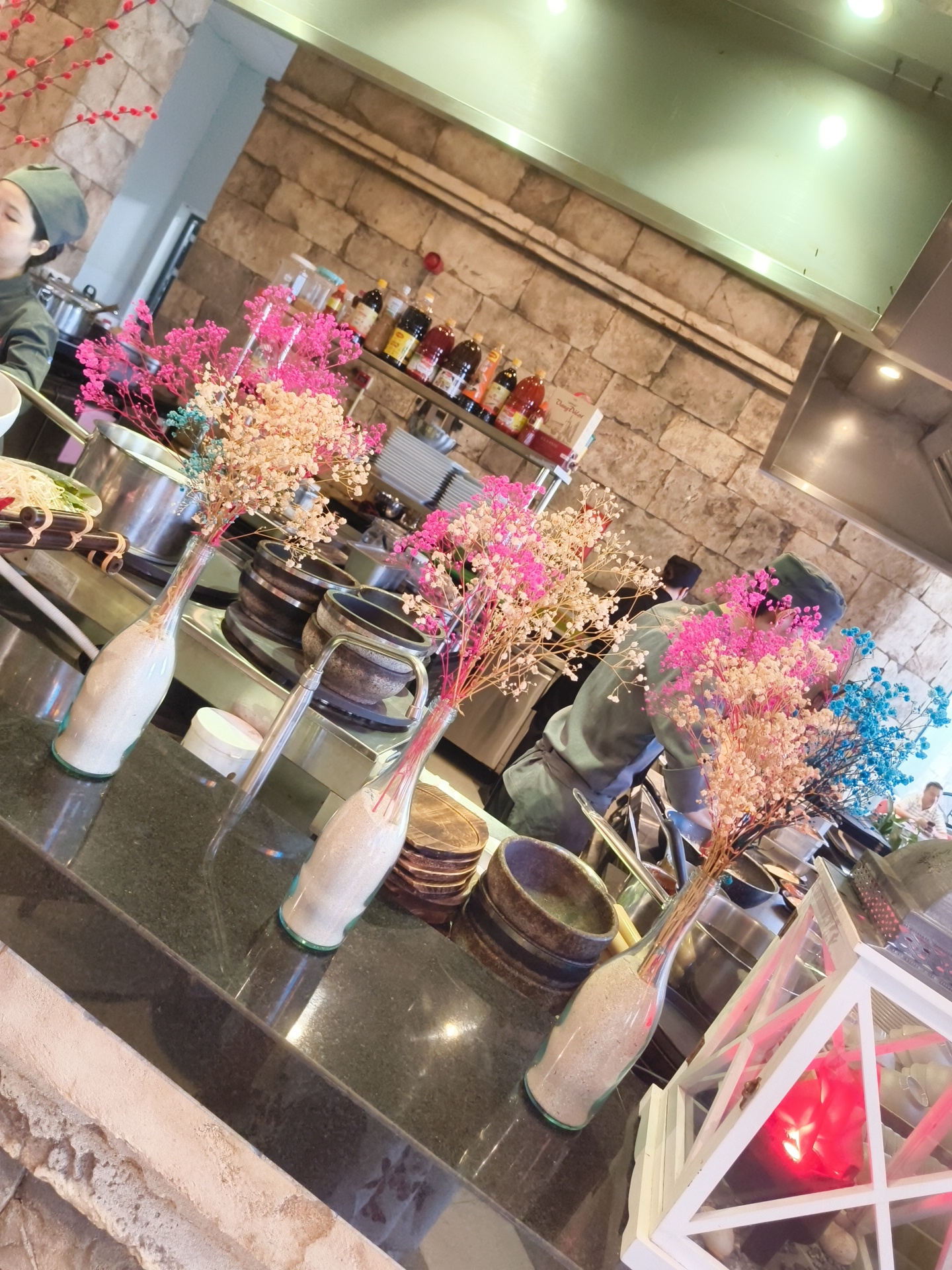 The image depicts a brightly lit interior scene, likely a restaurant kitchen or dining area. In the foreground, several glass vases filled with sand and topped with colorful dried flowers (pink, beige, and blue) are arranged on a dark, polished countertop. Behind the flowers, various cooking pots and bowls are visible on stovetop burners. A chef wearing a green hat and apron is seen in the middle ground, working at the counter. In the background, shelves are stocked with bottles of sauces and condiments, and a stone-clad wall forms the backdrop. To the right, a white decorative lantern with a red light inside is positioned on the counter. The overall impression is one of a clean, organized, and potentially upscale establishment, possibly located in Phú Quốc, Vietnam, given the context provided.