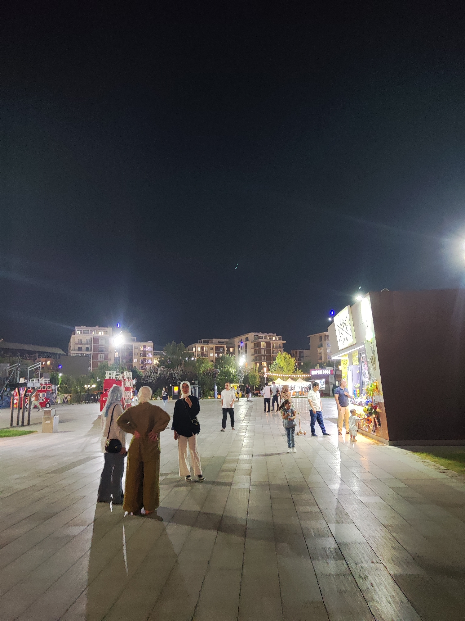 The image depicts an outdoor night scene in a modern urban area. In the foreground, a wide, illuminated tiled pathway reflects the light. Several people are present, walking and standing. To the left, there are some red structures that appear to be part of a children's play area or themed attraction, resembling stylized fire trucks.
Further back, a row of buildings with multiple floors and illuminated windows lines the background. These buildings have a contemporary architectural style. Trees are interspersed between the buildings and the pathway. On the right side of the image, a brightly lit kiosk or stall is visible, displaying various items, suggesting a commercial area or marketplace.
The lighting is characteristic of a city at night, with artificial lights from buildings, the pathway, and the stall creating a well-lit environment, contrasting with the dark sky. The overall impression is of a lively public space, possibly a park or plaza, in the city of Tashkent, Uzbekistan, known for its modern developments and public spaces.