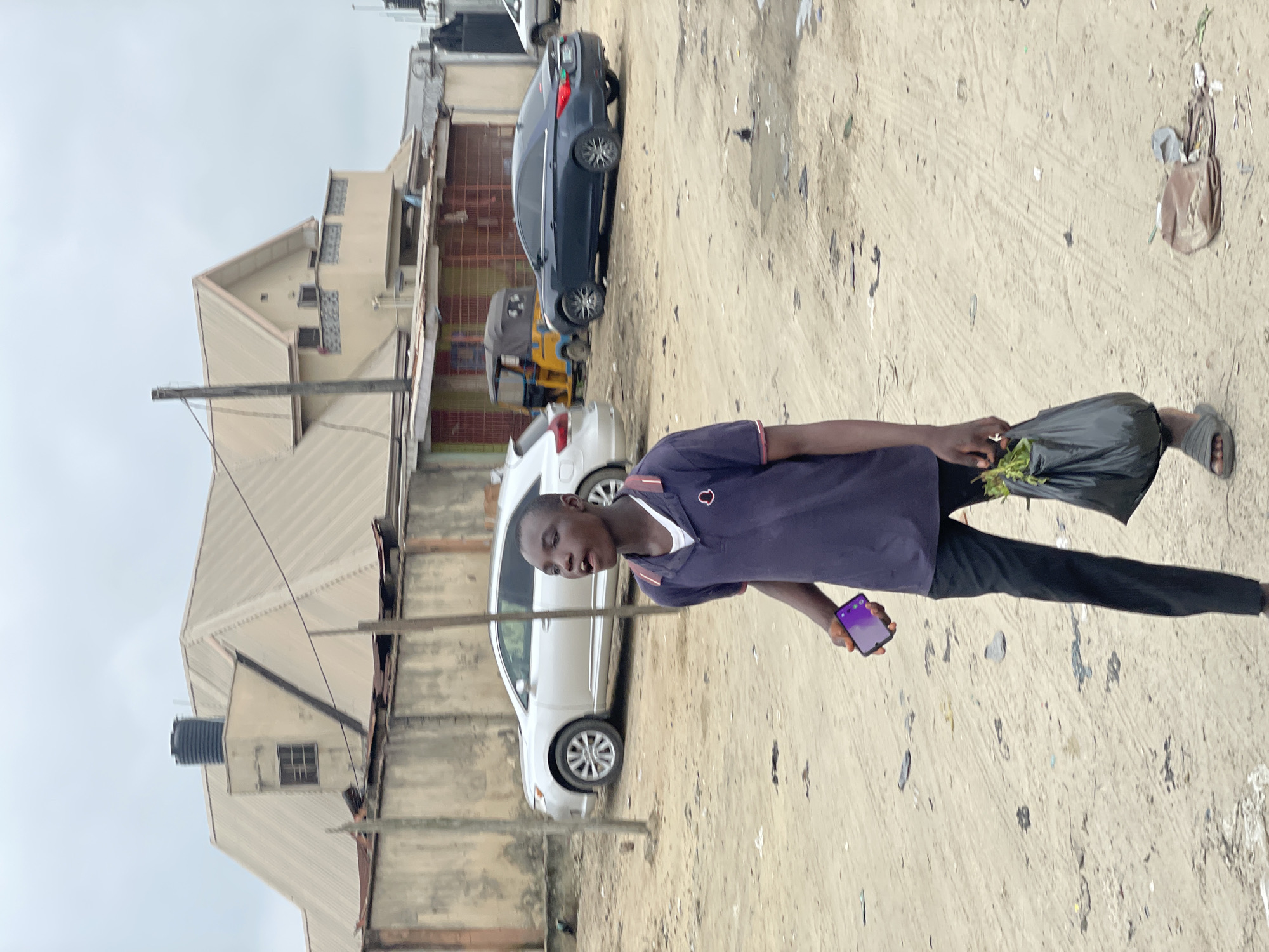 In Ado, Nigeria, a young Black male walks across a dusty, unpaved area under an overcast sky. He is wearing a dark blue polo shirt with a white collar, black trousers, and grey slide sandals. In his left hand, he holds a dark plastic bag filled with green leafy vegetables. In his right hand, he carries a purple smartphone. Behind him, a silver SUV is parked on the dirt road. Further in the background, a yellow and black three-wheeled auto-rickshaw is visible. Several buildings with light-colored, sloped roofs and concrete walls are present in the distance. The ground is covered in sand and scattered debris.