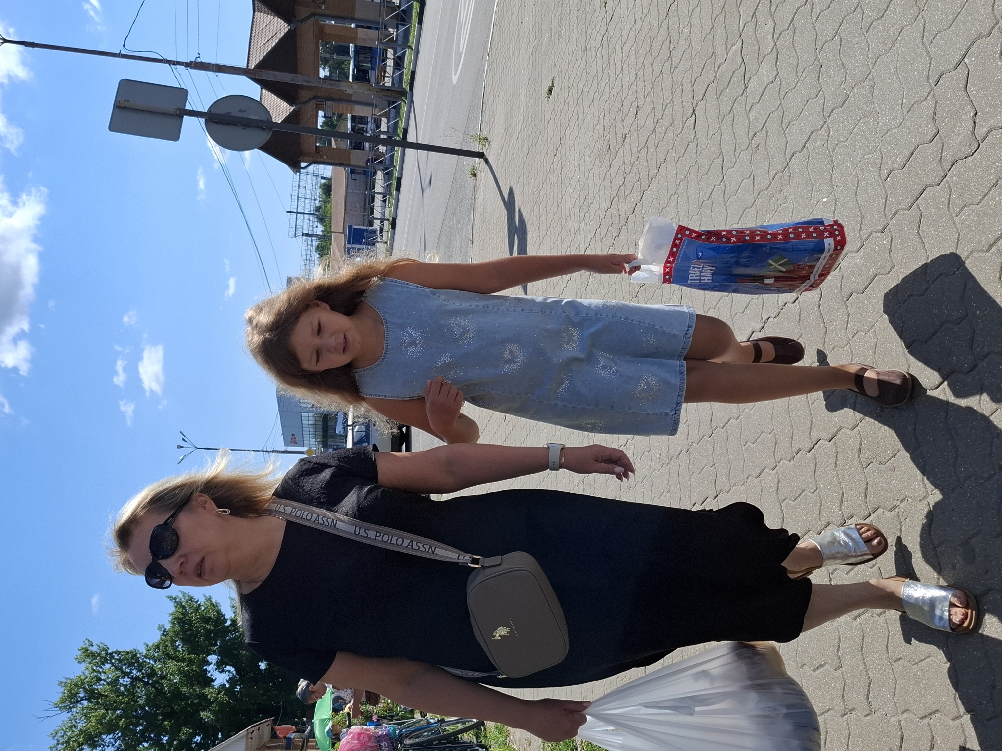 A medium shot shows a woman and a young girl walking on a sidewalk under a bright, cloudy sky. The woman, on the left, wears a black dress, sunglasses, and silver sandals. She carries a large white plastic bag in her left hand and a dark gray crossbody purse with a U.S. Polo Assn. strap. The girl, on the right, wears a light blue denim dress adorned with rhinestones and brown Mary Jane shoes. She holds a colorful shopping bag with images of cartoon characters and text. They appear to be walking away from the viewer, casting distinct shadows on the patterned concrete sidewalk. In the background, there's a street with a road and a few buildings that resemble bus stops or small shops. Trees are visible on the far left, and a street sign stands on a pole to the right. The overall scene suggests a sunny day in a public outdoor space.