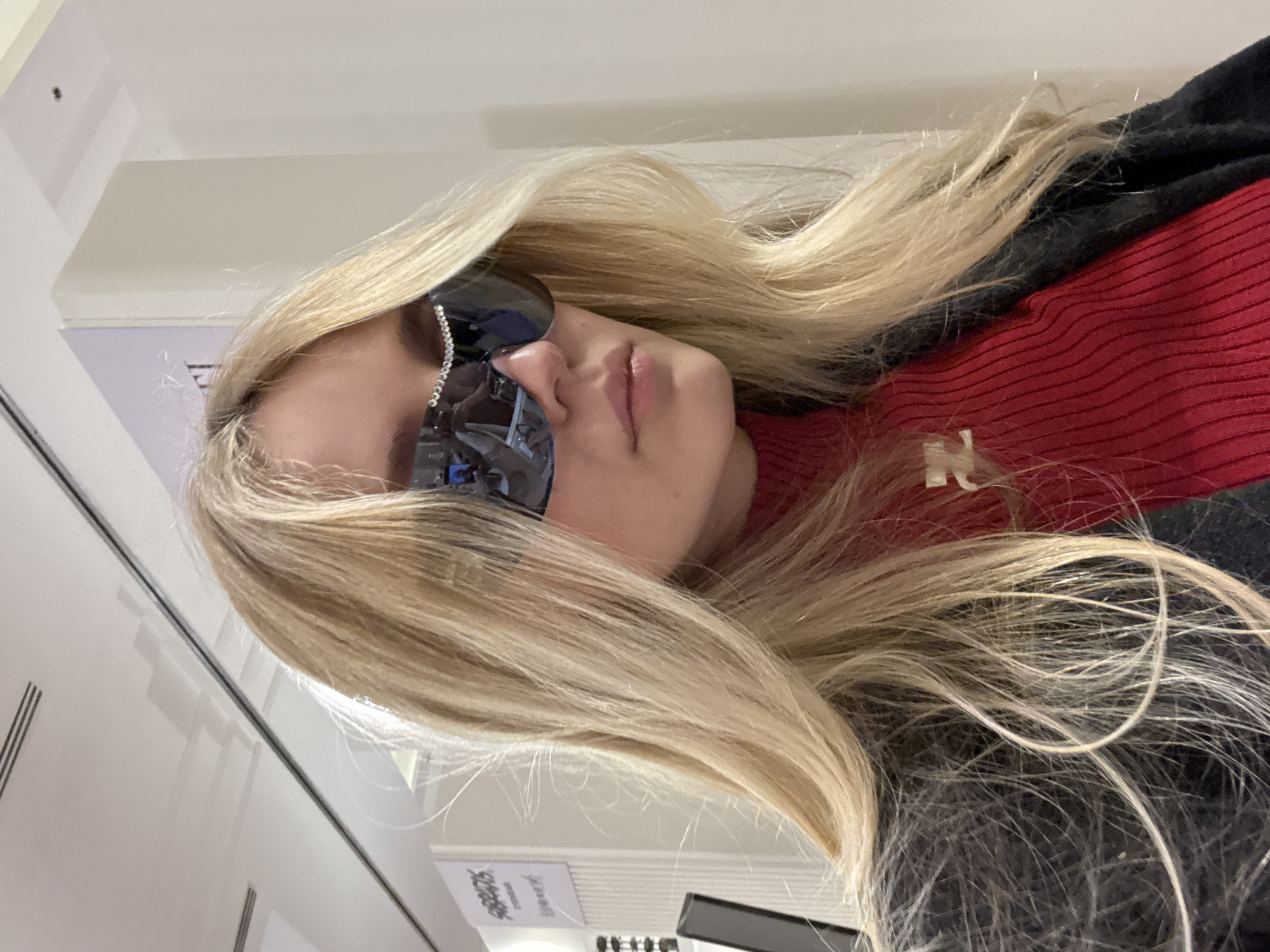 A woman with long blonde hair is wearing sunglasses with rhinestone embellishments. She is wearing a red sweater with a dark-colored top layer, likely a jacket or coat. The environment suggests an indoor setting, possibly a store or commercial space with reflective surfaces. The location in Mexico City is not discernible based on the visible elements in the image.