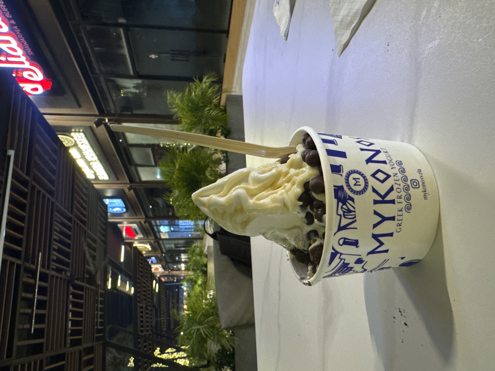 The image shows a cup of Mykonos Greek frozen yogurt topped with chocolate chips, with a wooden spoon inserted. The background depicts a nighttime street scene, with buildings and trees illuminated with yellow lights. The text "shawarma & buffet" is visible on a red sign on one of the buildings. The location is Pampatar, Venezuela.